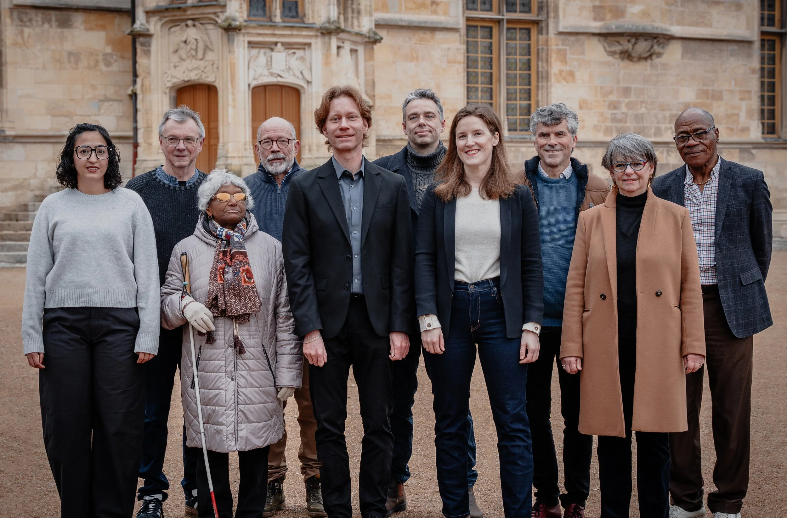 Groupe de dix personnes posant devant un bâtiment historique en pierre, certains portant des vêtements d'extérieur et des lunettes, souriant à la caméra.