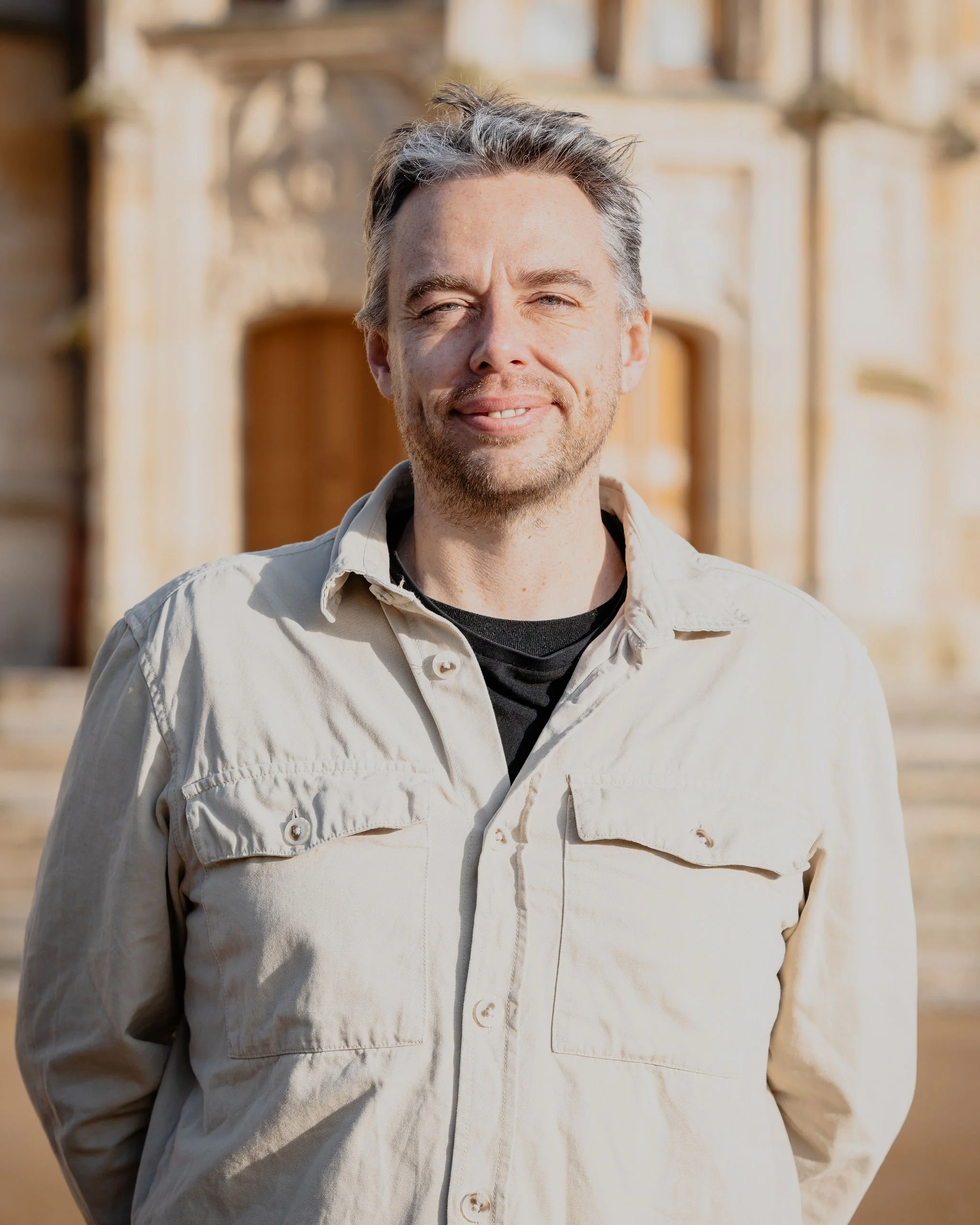 Un homme souriant porte une veste beige, se tenant devant un bâtiment ancien en pierre.