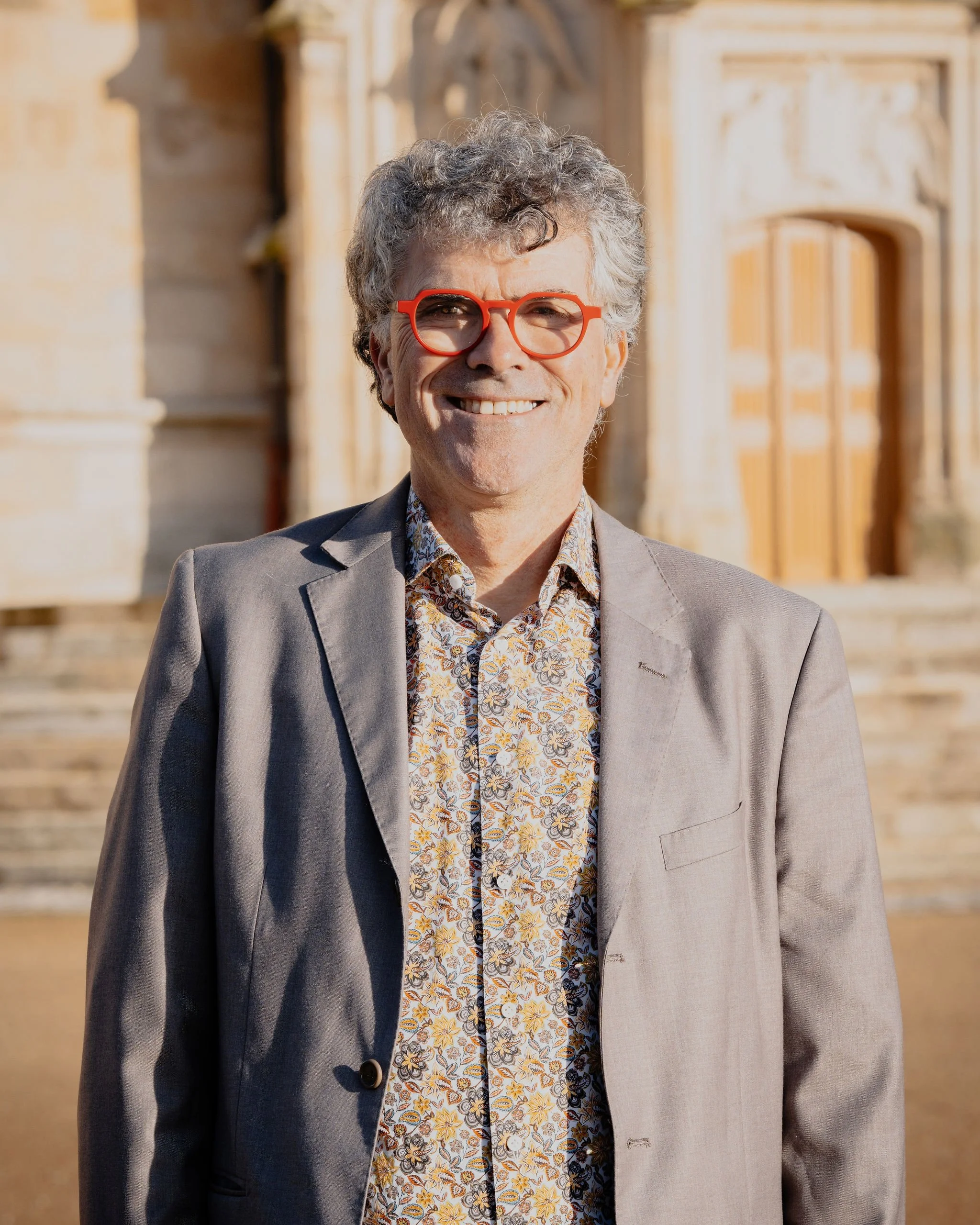 Homme souriant portant des lunettes rouges, costume gris, chemise à motifs floraux, devant un bâtiment en pierre avec des escaliers.