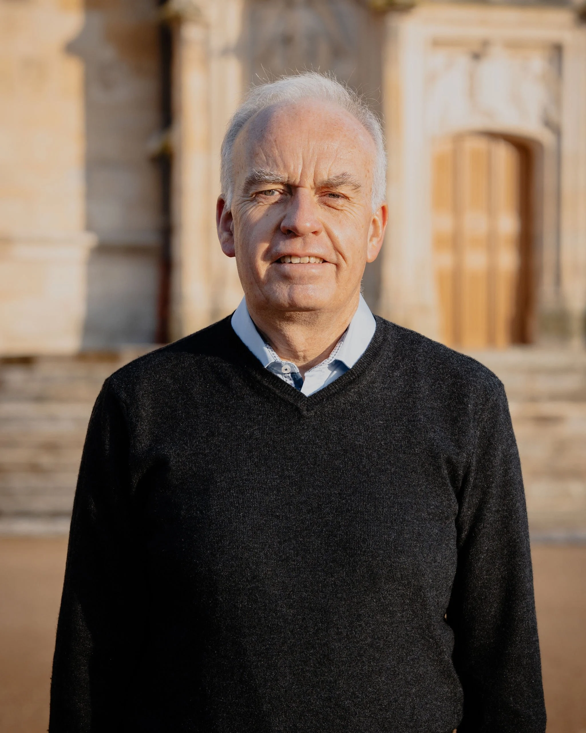 Un homme âgé dehors, portant un pull noir et une chemise blanche, devant une vieille bâtisse en pierre, ensoleillée.