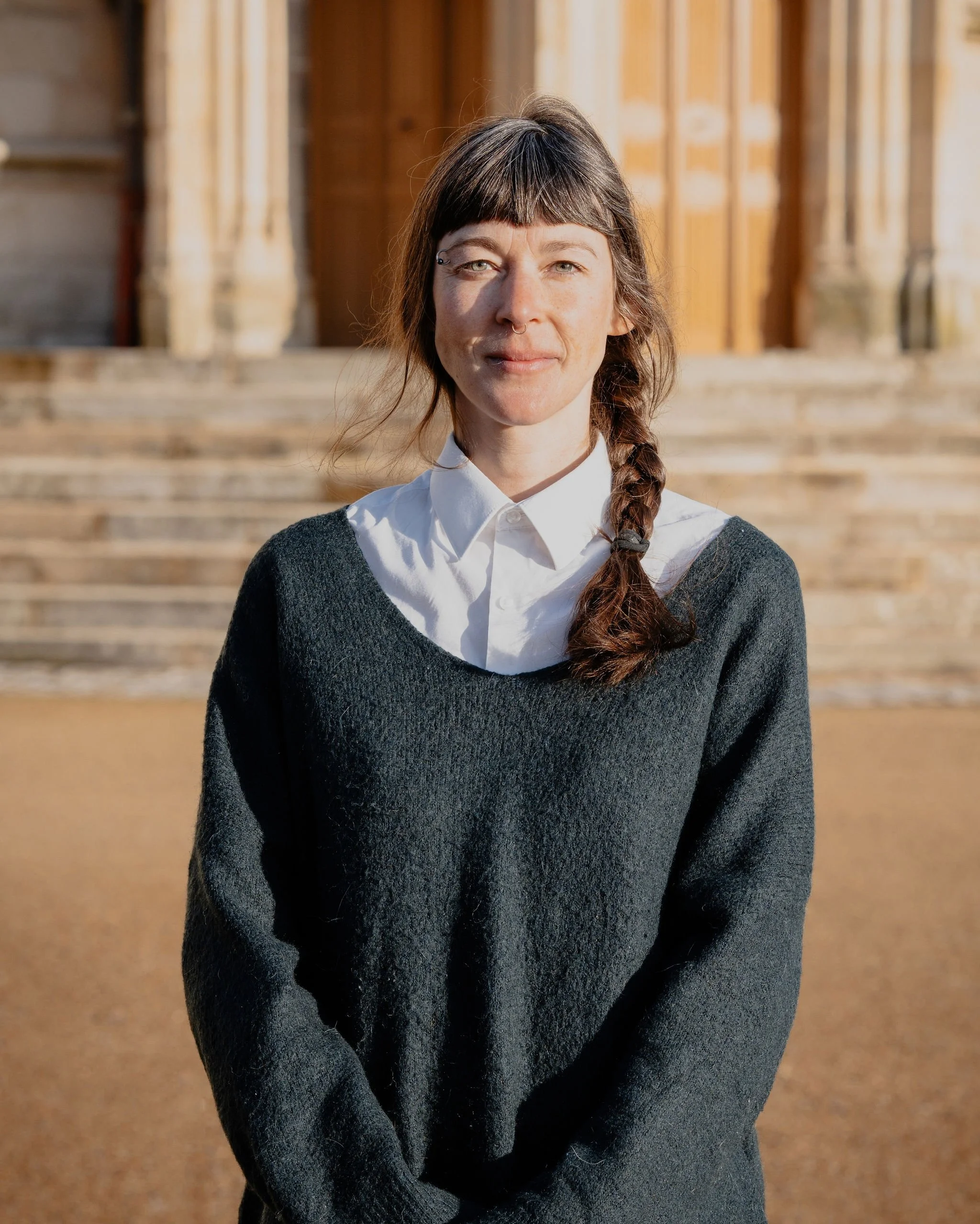 Une femme debout devant une marche en pierre, portant une chemise blanche et un pull gris foncé, avec un bâtiment en pierre en arrière-plan.