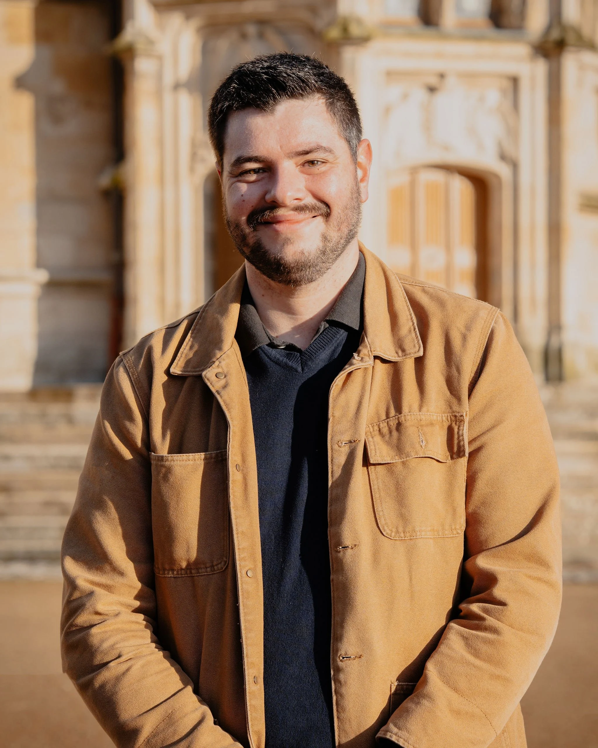 Jeune homme souriant portant une veste marron, devant un bâtiment ancien en pierre, en plein jour, avec des escaliers en pierre en arrière-plan.