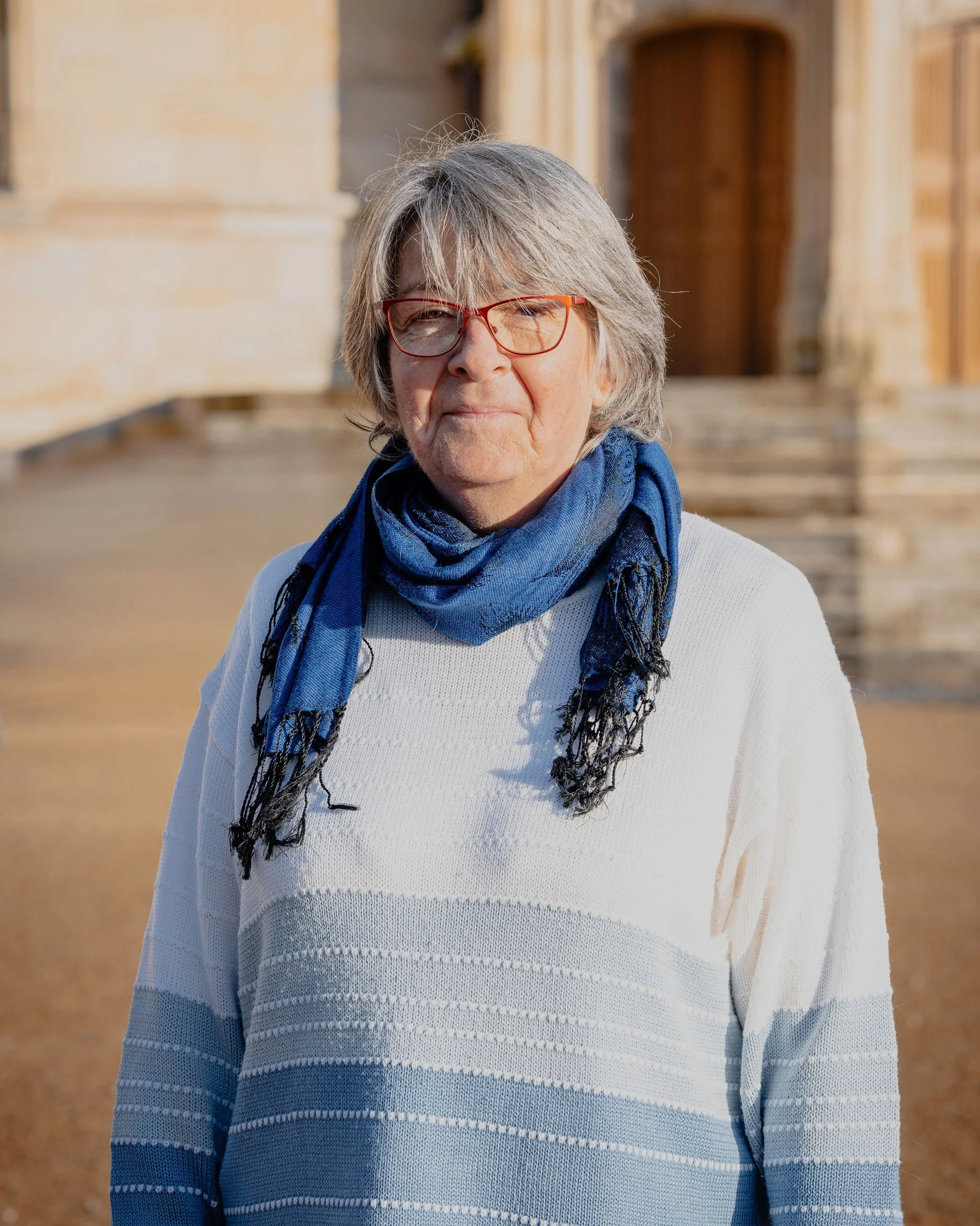Une femme âgée avec des lunettes rouges, un foulard bleu et un pull blanc, debout devant une église en pierre, dans un endroit ensoleillé.