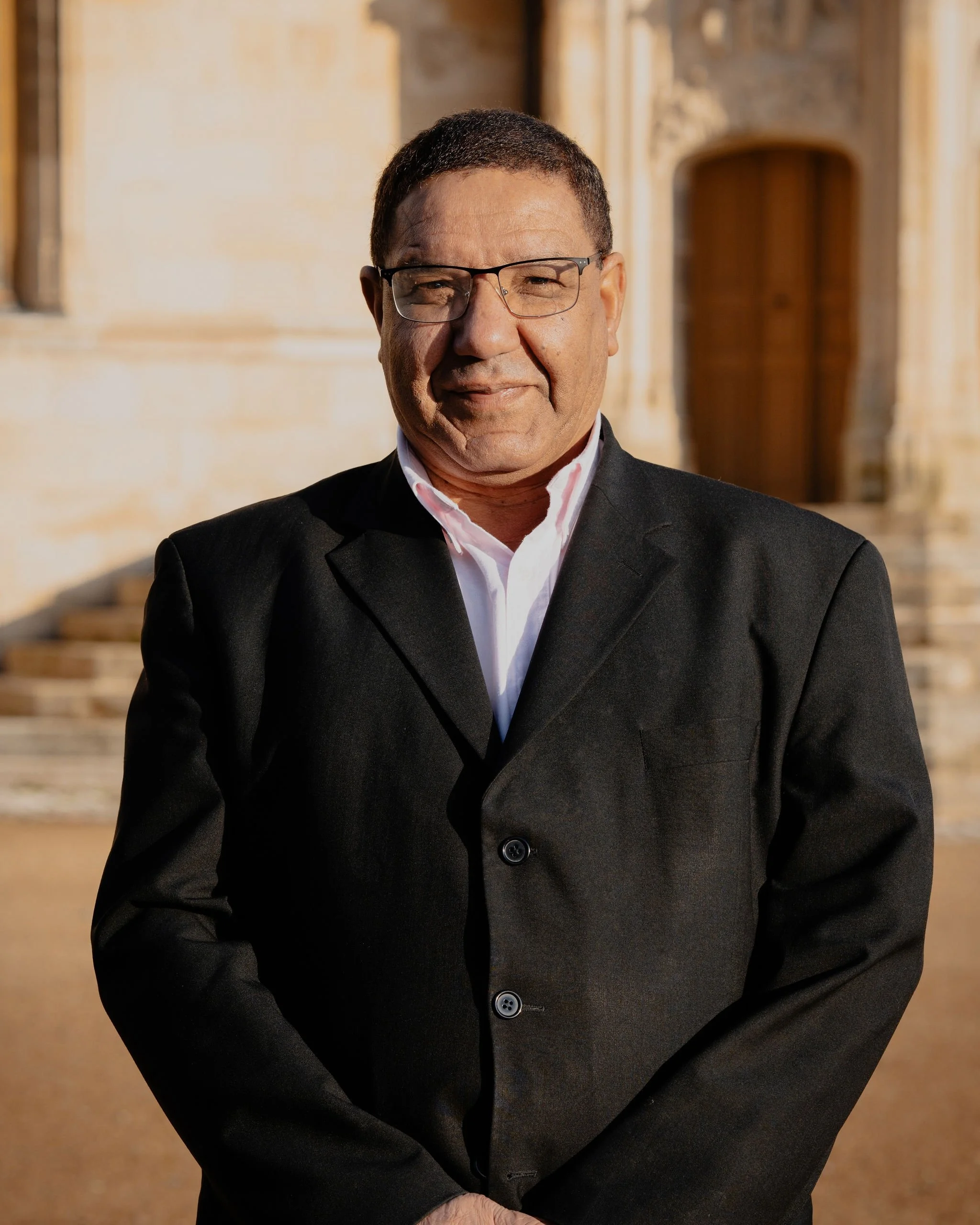 Un homme en costume noir avec une chemise rose pâle, portant des lunettes, se tient devant un bâtiment en pierre au crépuscule.
