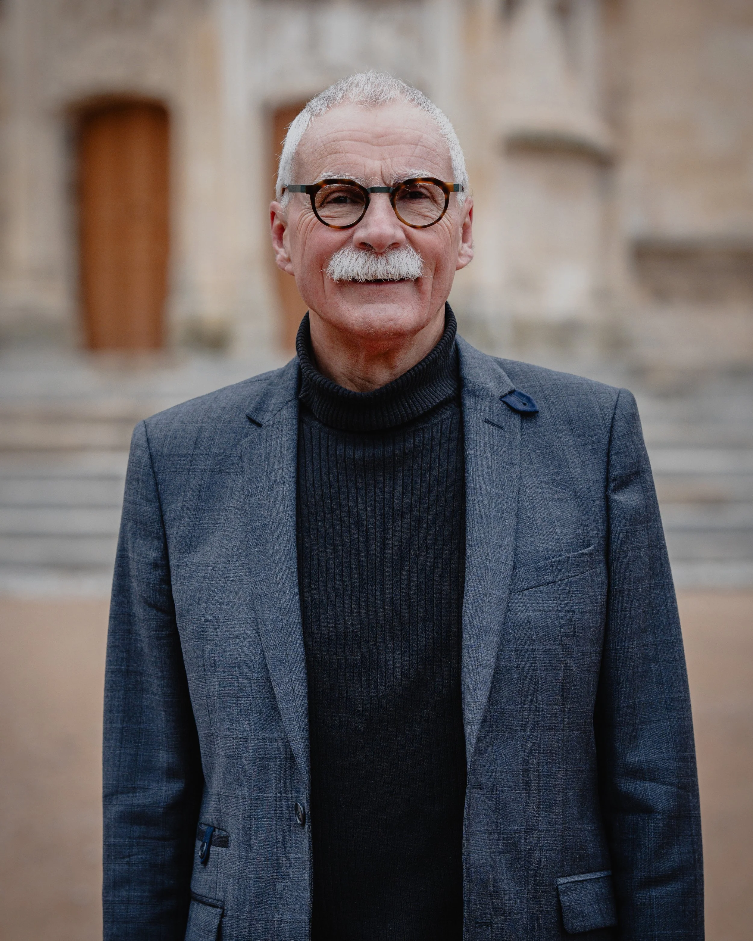 Un homme âgé avec des lunettes rondes, une moustache blanche, portant un costume gris avec un pull noir, se tenant devant un bâtiment ancien en pierre.