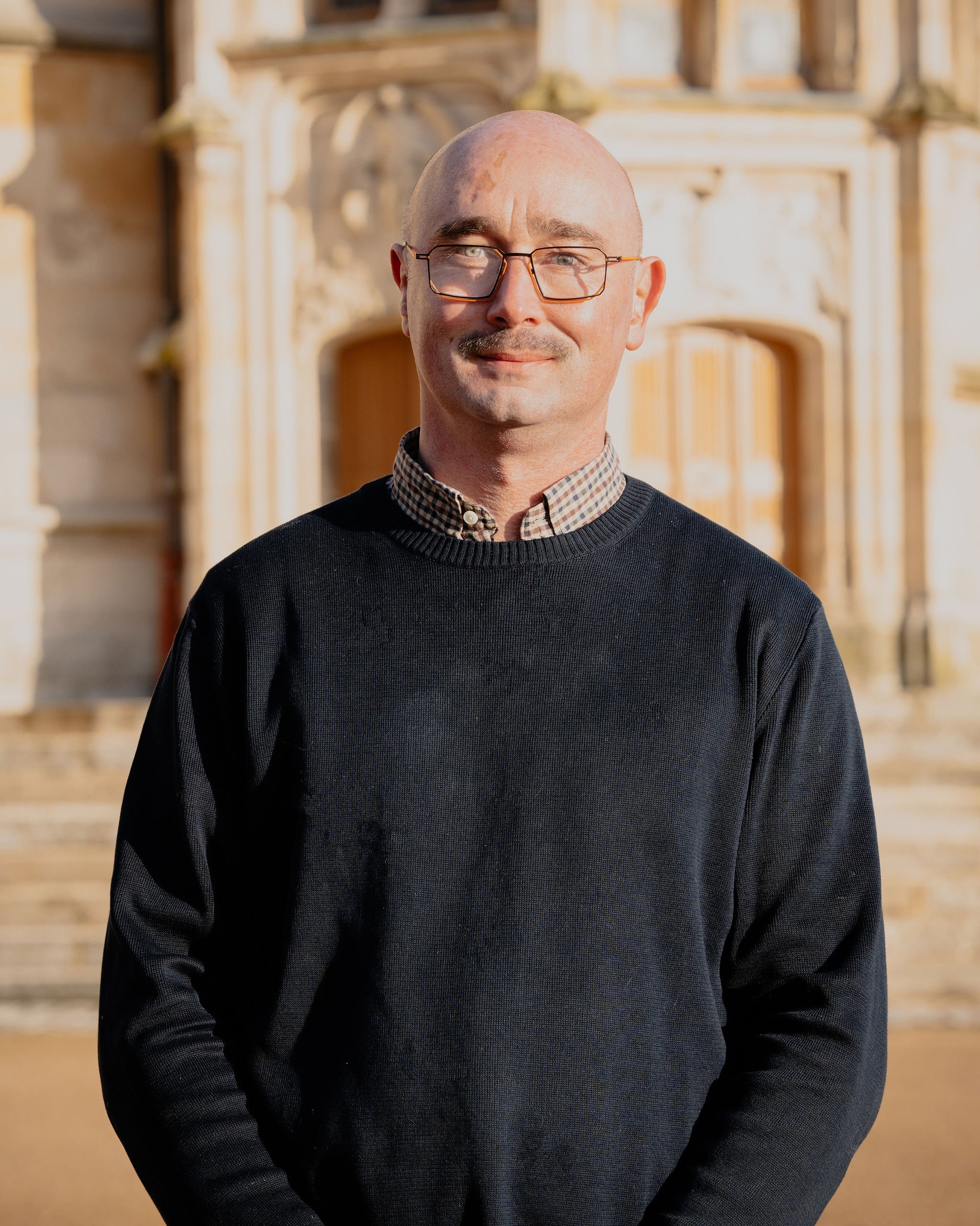 Un homme souriant portant des lunettes, un pull noir et une chemise à carreaux, se tenant devant un bâtiment en pierre ancienne, en plein jour.