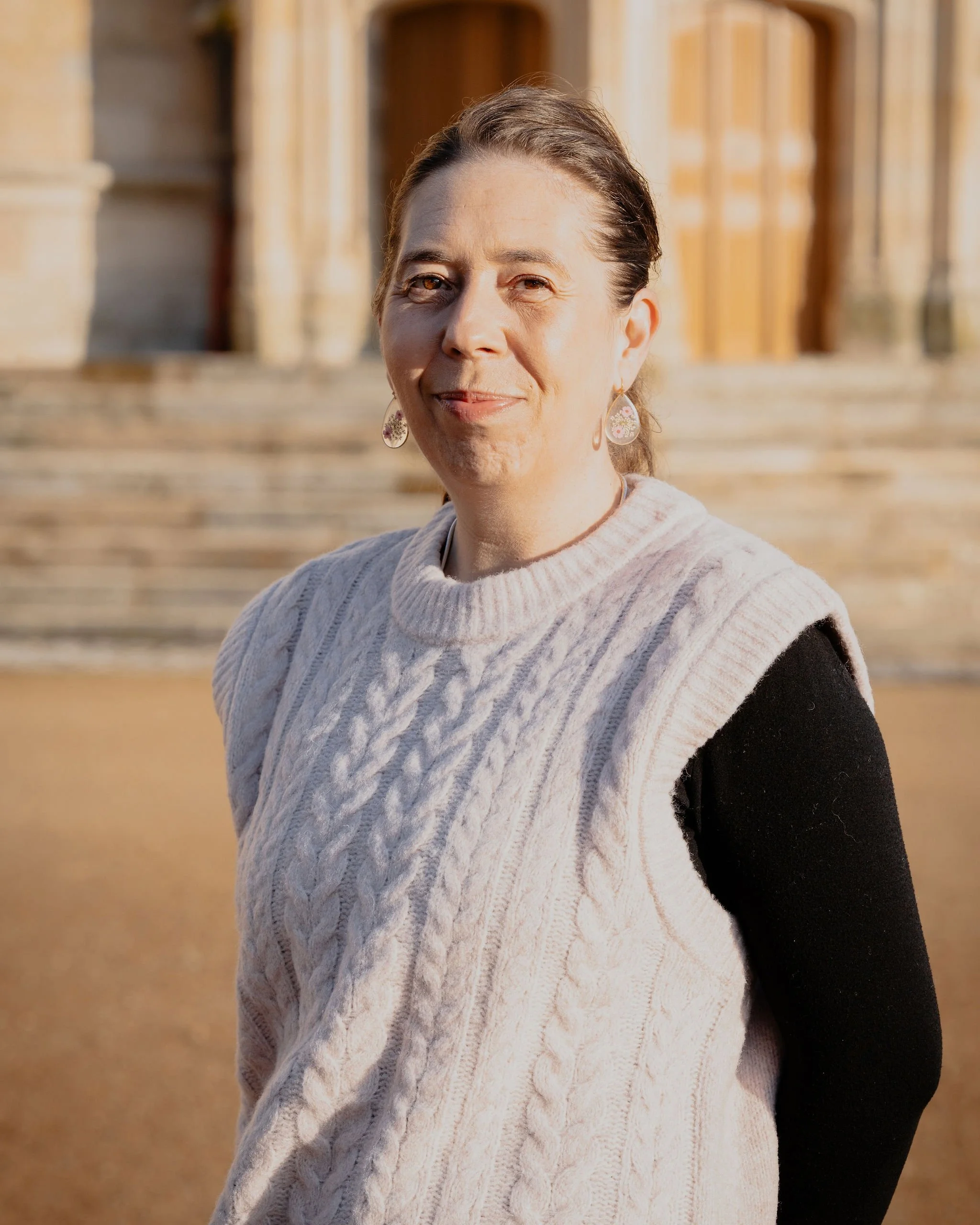 Une femme souriante portant un pull beige en laine et des boucles d'oreilles, avec des escaliers en pierre et un bâtiment ancien en arrière-plan.