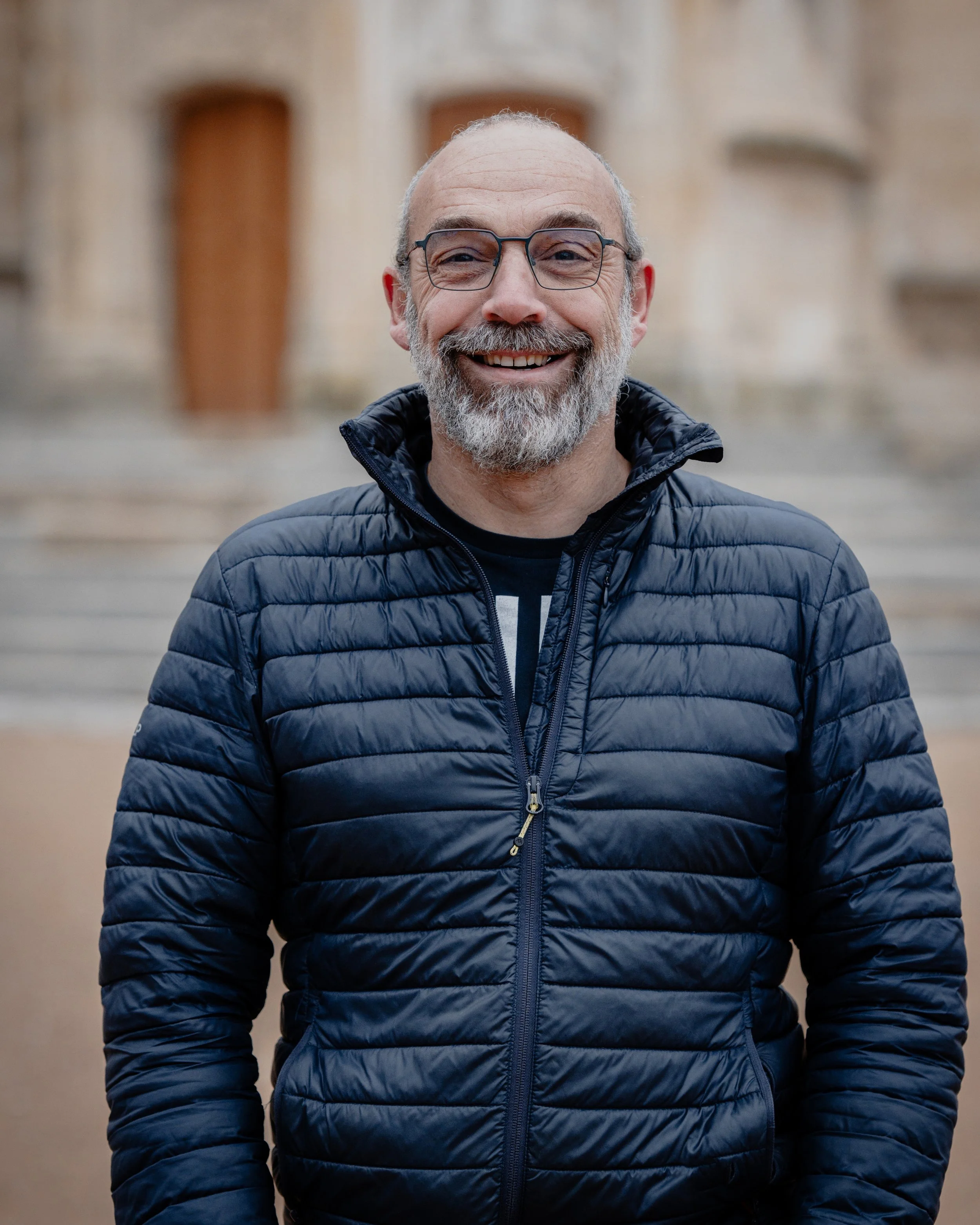 Homme souriant avec lunettes, barbe grise, portant une veste noire, devant un bâtiment ancien en pierre.
