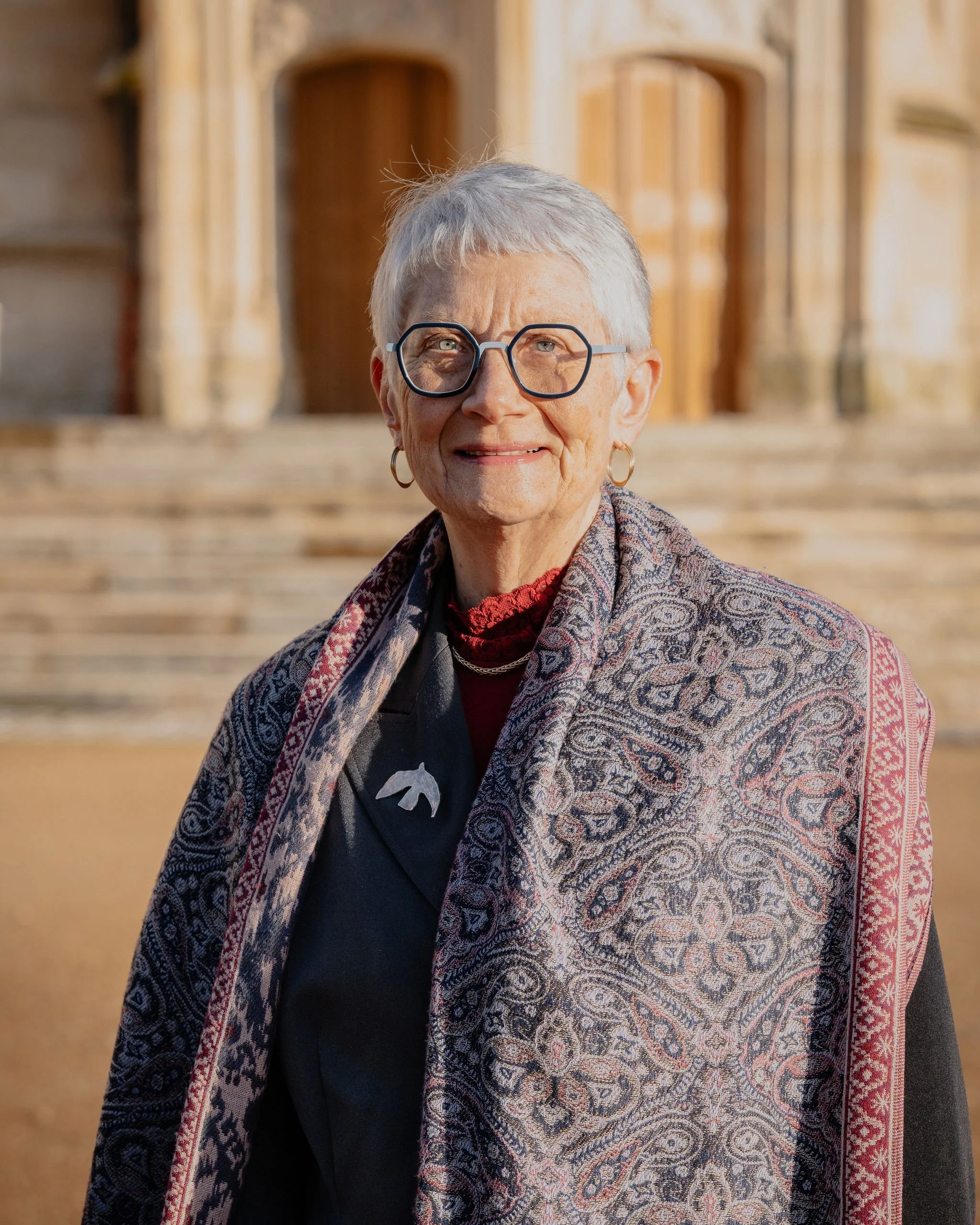 Portrait d'une femme âgée avec des cheveux gris, portant des lunettes, un costume noir et une écharpe colorée, debout devant un bâtiment en pierre.