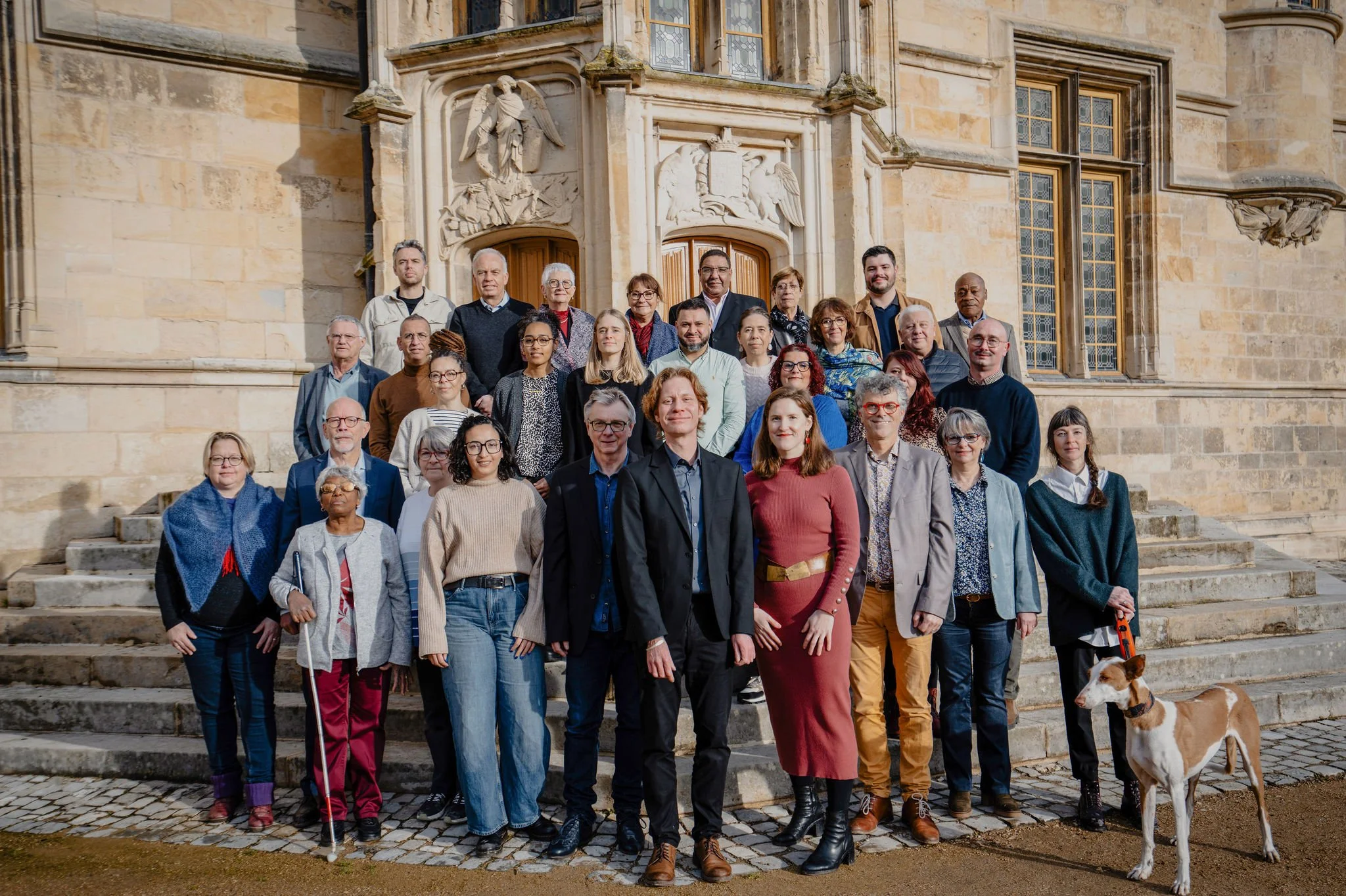 Groupe de personnes posant devant une vieille bâtiment en pierre, avec des fenêtres ornées et des sculptures, lors d'une sortie en extérieur, un chien est aussi présent.