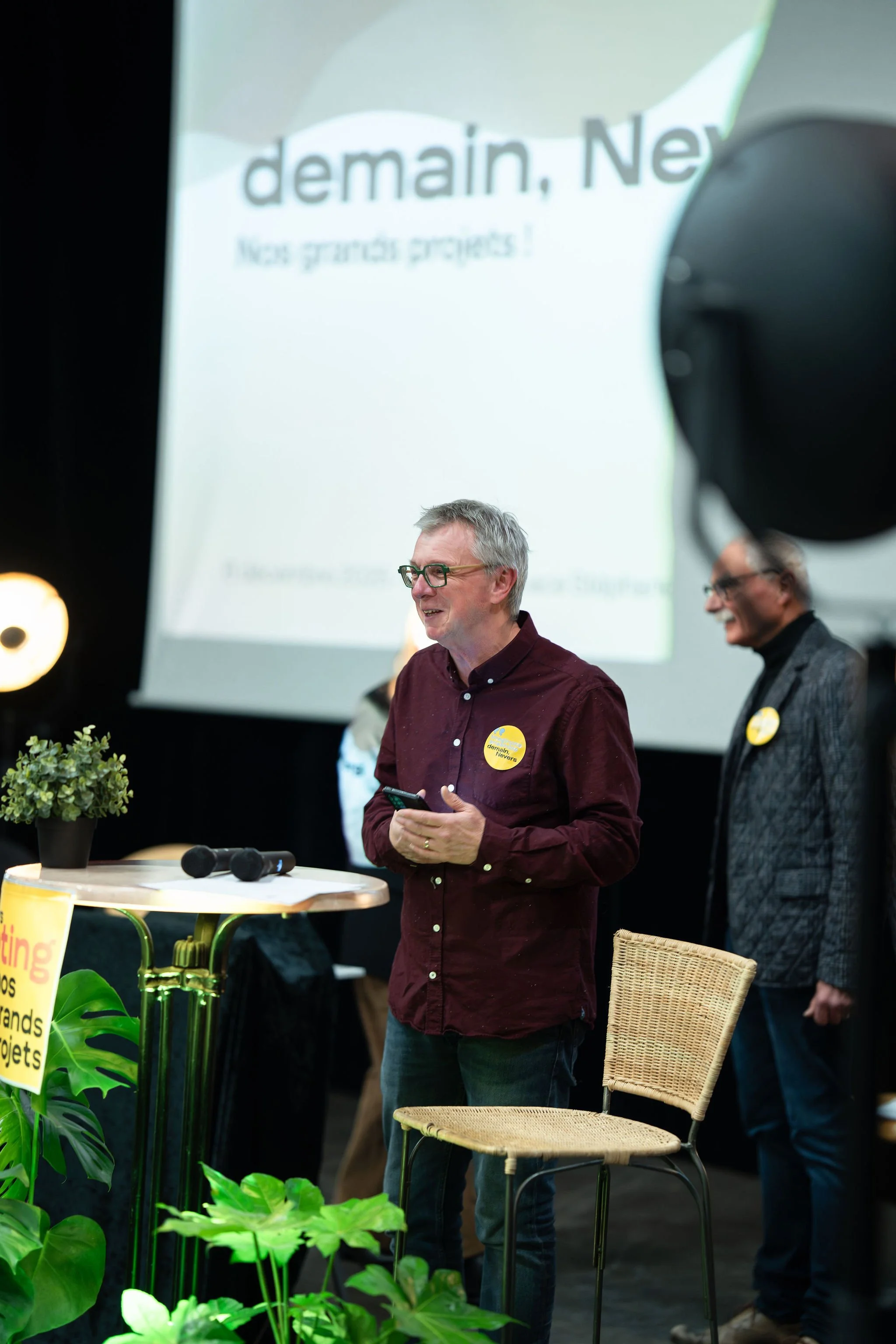 Deux hommes parlent lors d'une présentation ou d'un événement, avec un écran en arrière-plan affichant le mot 'demain, Ne...' et des plantes en premier plan.