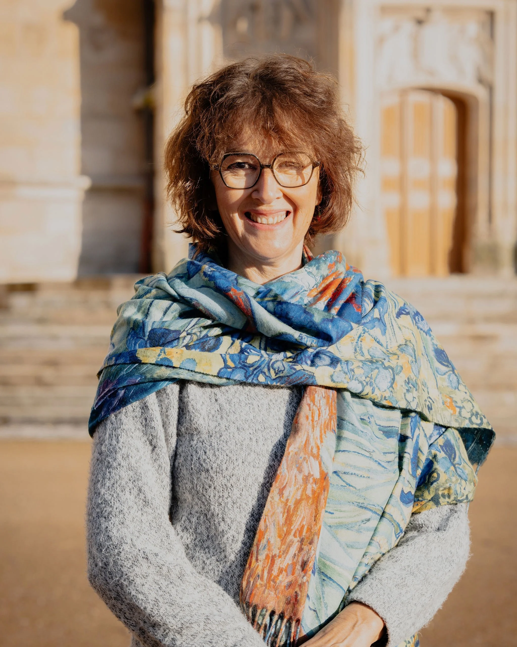 Femme souriante portant des lunettes, un foulard coloré et un manteau gris, en extérieur avec un bâtiment ancien en arrière-plan.