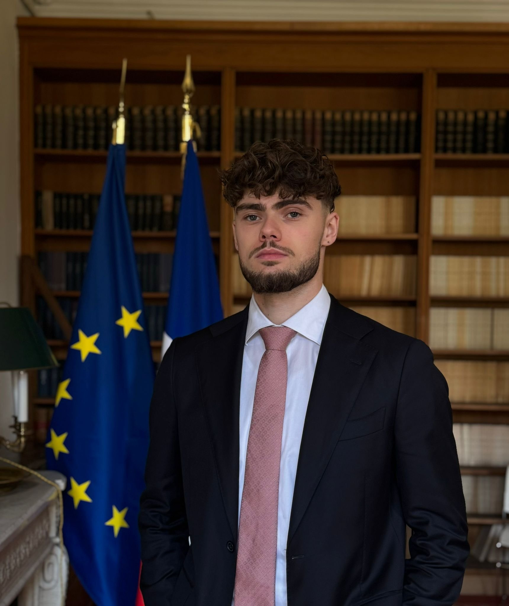 Jeune homme en costume dans une salle avec des drapeaux de l'Union européenne et de la France en fond, bibliothèque en arrière-plan.