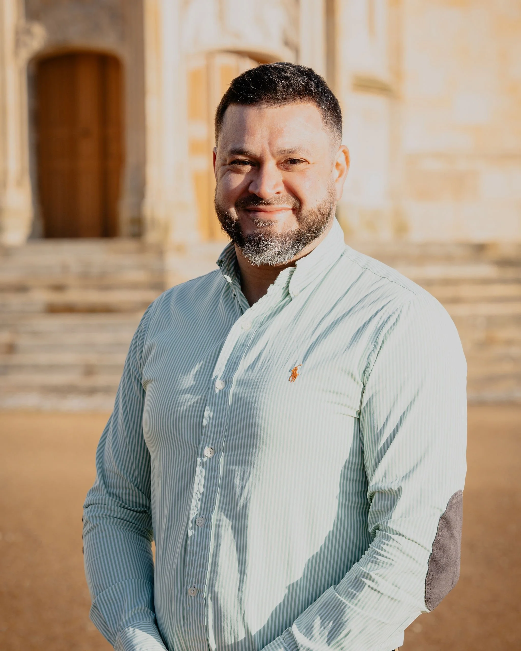 Un homme souriant portant une chemise à rayures avec un bâtiment ancien en pierre en arrière-plan.