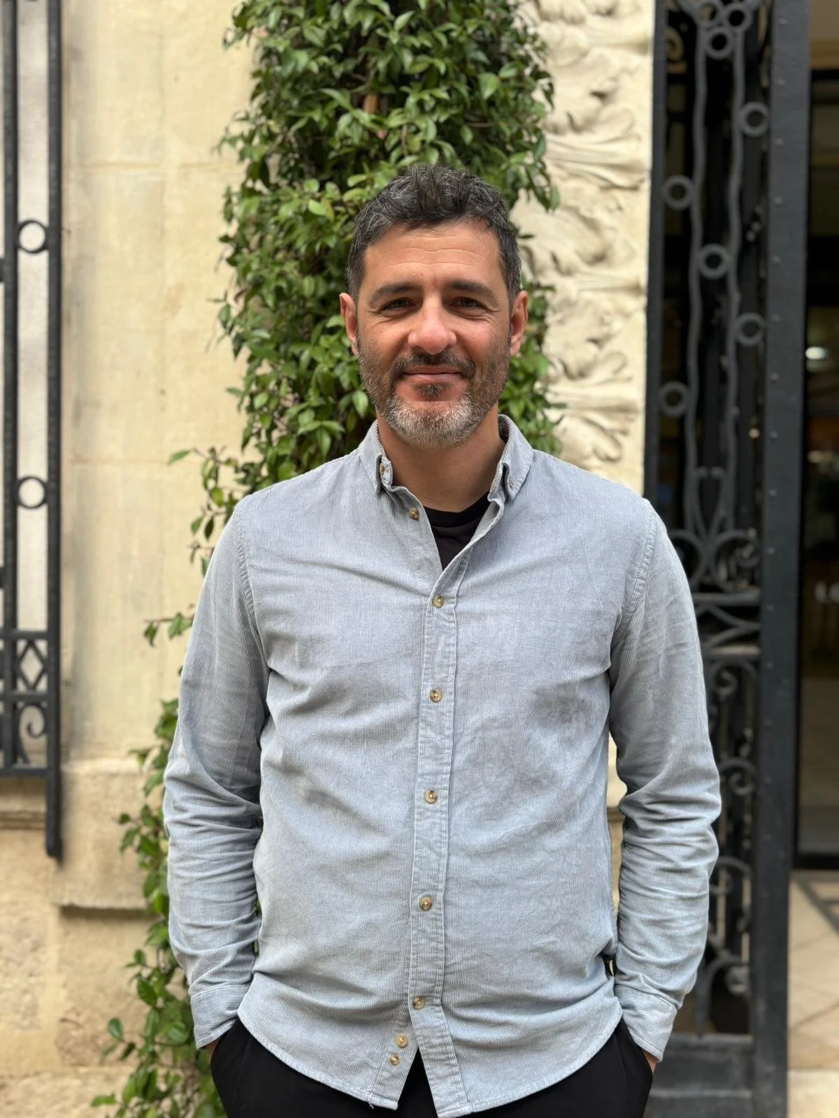 Un homme souriant, cheveux noirs, barbe grise, portant une chemise grise décontractée, posé devant un mur avec de la végétation et une grille en fer noir.