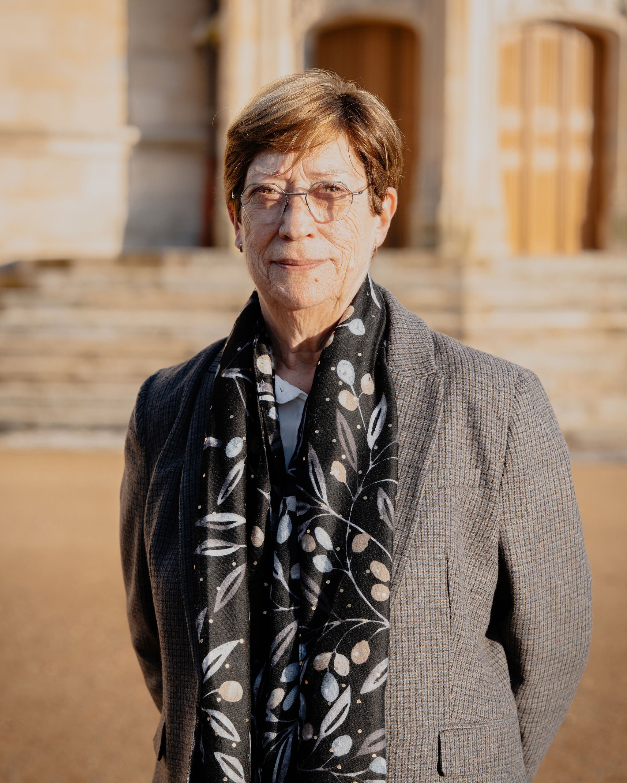 Une femme âgée portant une veste à carreaux, une écharpe à motifs de feuilles, et des lunettes, posant devant un bâtiment en pierre avec un escalier en arrière-plan.