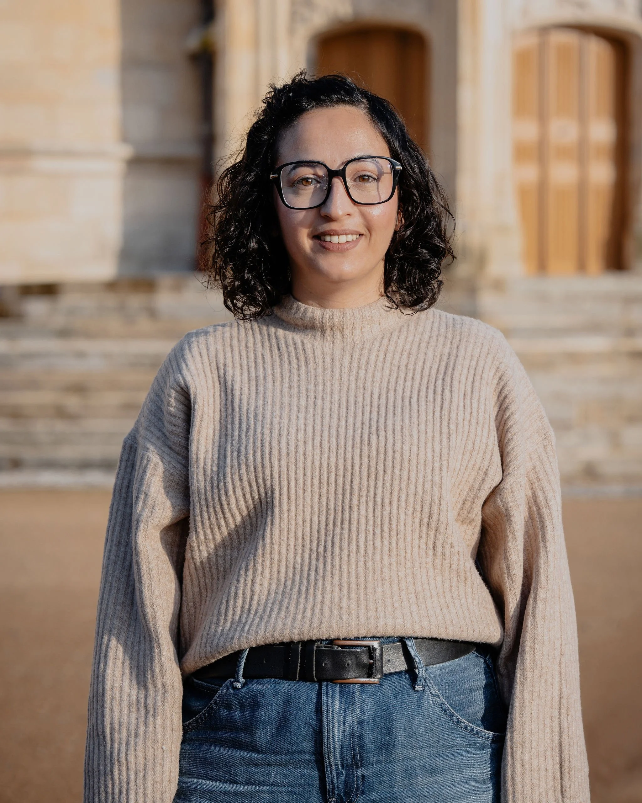 Jeune femme avec lunettes, portant un pull beige et un jean, devant un bâtiment en pierre.