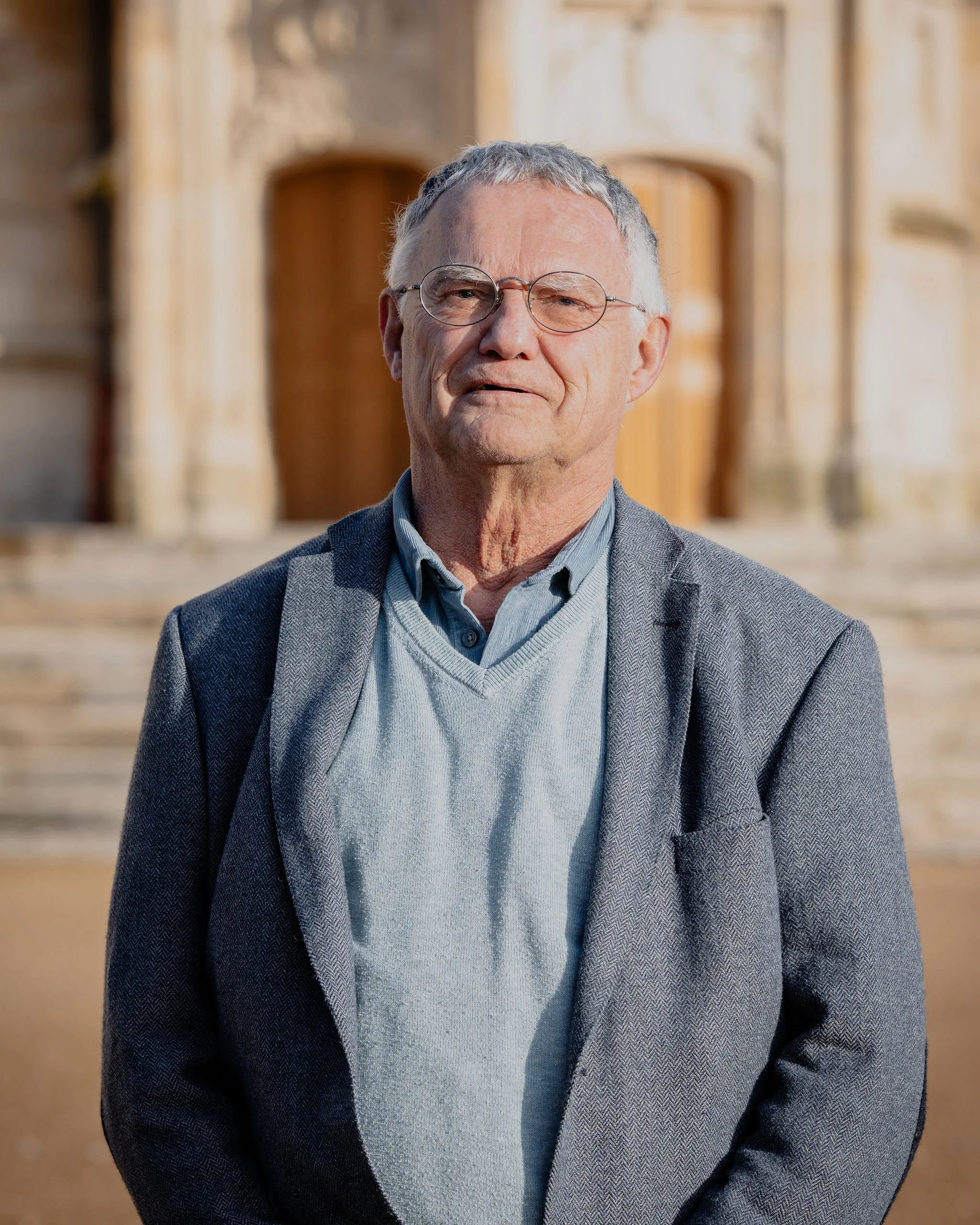 Un homme âgé portant des lunettes, une veste grise, un pull bleu clair et une chemise, se tient devant une ancienne église en pierre, probablement en France.