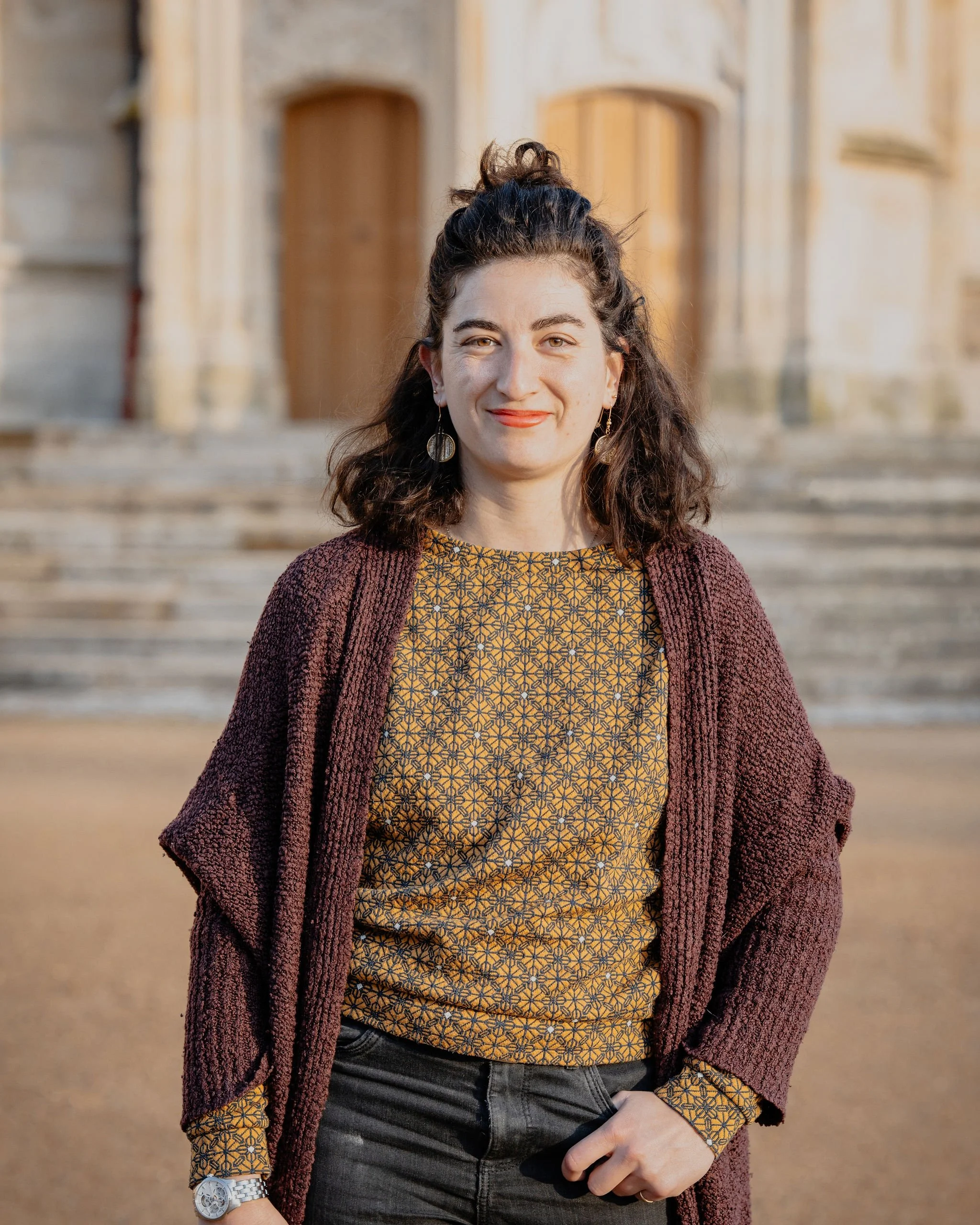 Une femme souriante avec des cheveux bouclés, portant un pull à motifs jaunes et bleus, une veste marron foncé et des boucles d'oreilles, devant un bâtiment en pierre avec des escaliers.