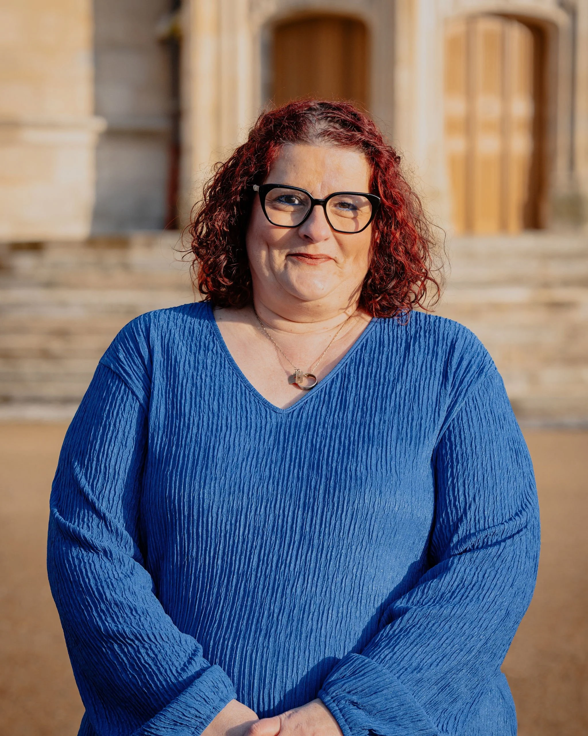Une femme aux cheveux bouclés rouges portant des lunettes noires, un pull bleu, debout devant un bâtiment en pierre avec des escaliers.
