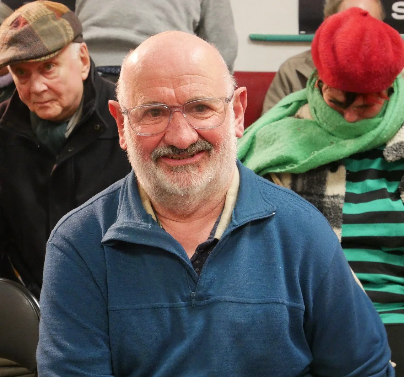Un homme âgé souriant avec des lunettes, portant une veste bleue, assis lors d'un rassemblement social, entouré de personnes.