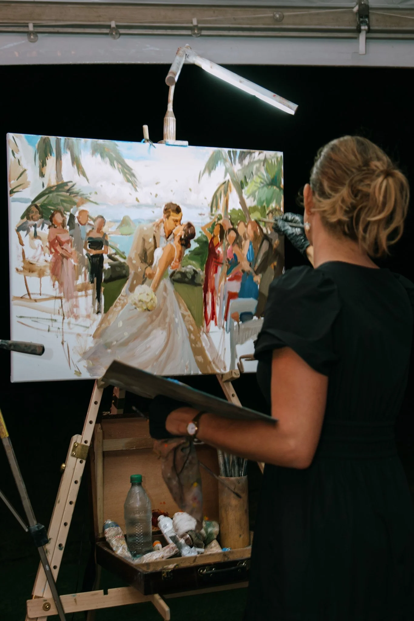 Artist painting a wedding scene on a canvas at an easel, depicting a couple kissing at a tropical location with onlookers. The artist stands in the foreground with art supplies nearby.