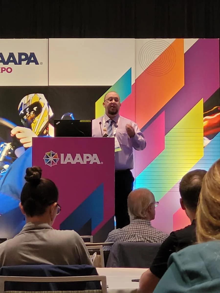 A man giving a presentation at the IAAPA Expo with a colorful background and a podium labeled 'IAAPA'. There are several audience members seated and listening.
