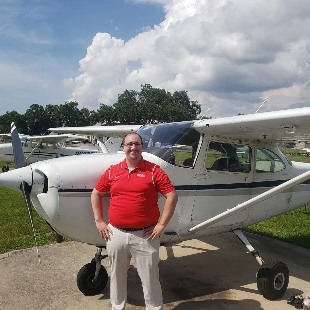A man in a red polo shirt and white pants standing in front of a small white airplane on a tarmac, with other airplanes and trees in the background under a partly cloudy sky.