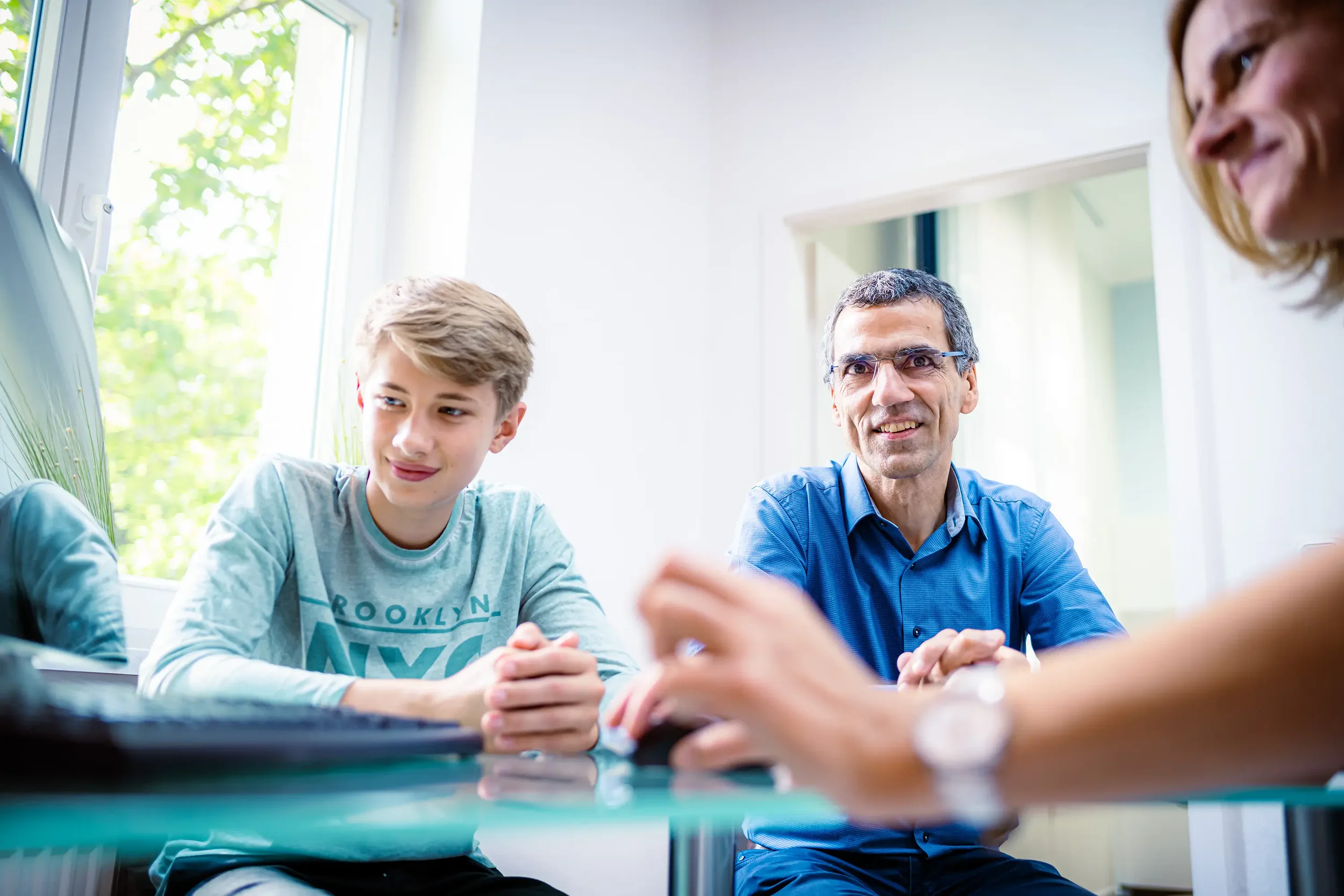Ein Junge und zwei Erwachsene sitzen an einem Tisch, sprechen und lächeln in einem hellen Raum mit großen Fenstern, die Tageslicht hereinlassen.