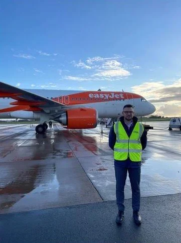 A man in a high-visibility vest standing in front of an easyJet airplane on an airport tarmac during daylight.