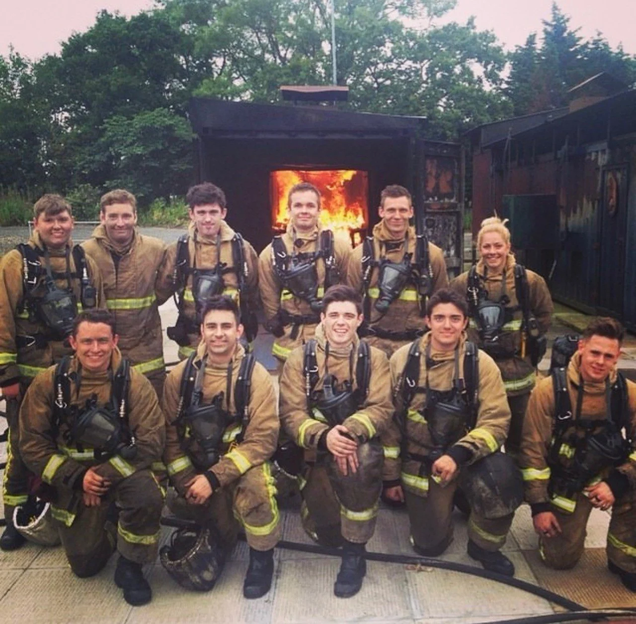 A group of eleven firefighters in uniform smiling on a fire station porch with an outdoor fire simulator in the background.