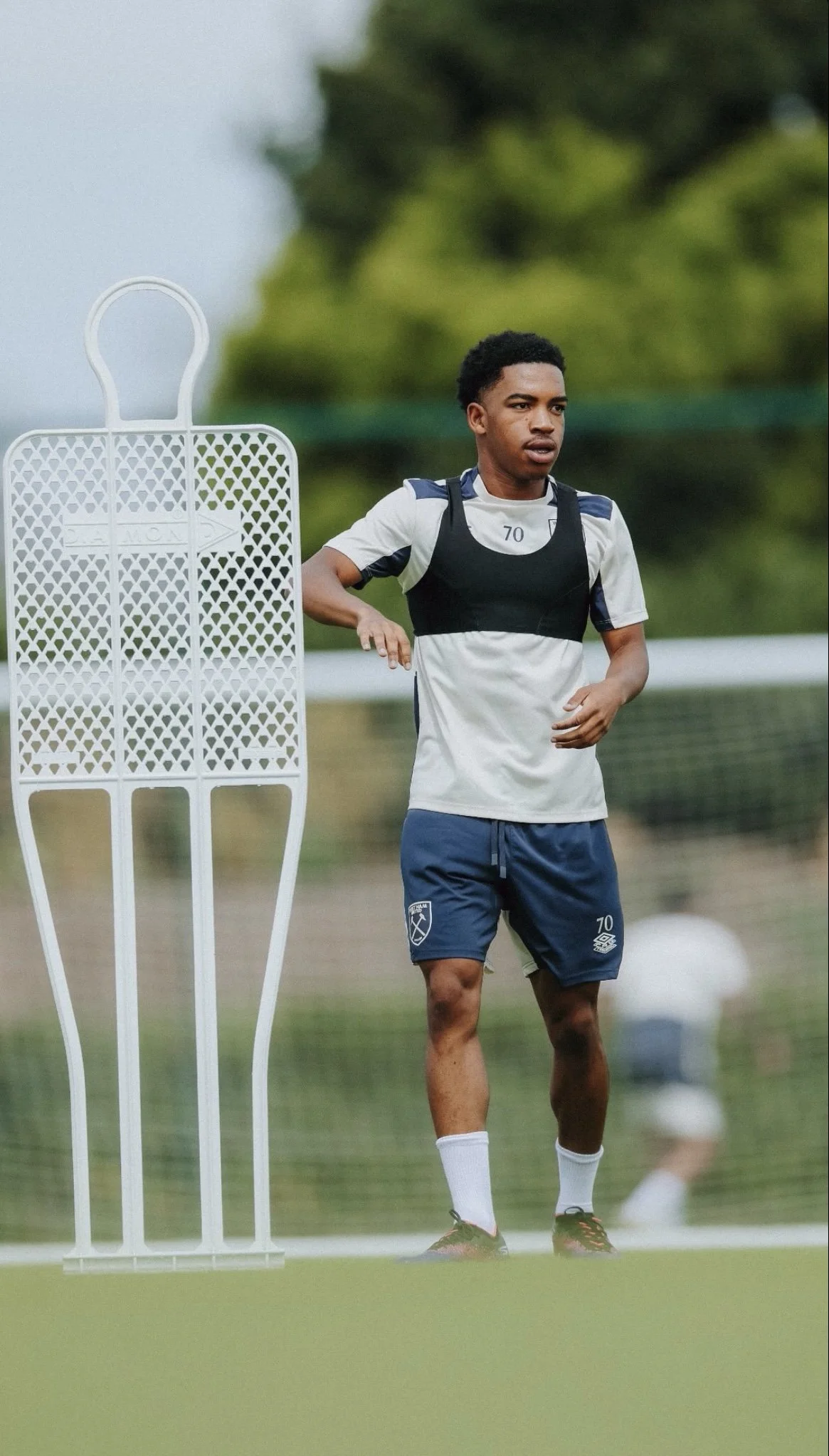 Elisha Sowunmi playing for West Ham United in a white and navy uniform with the number 70, wearing a training vest, standing on a soccer field during practice. There is a white training mannequin next to him and a goal net in the background.