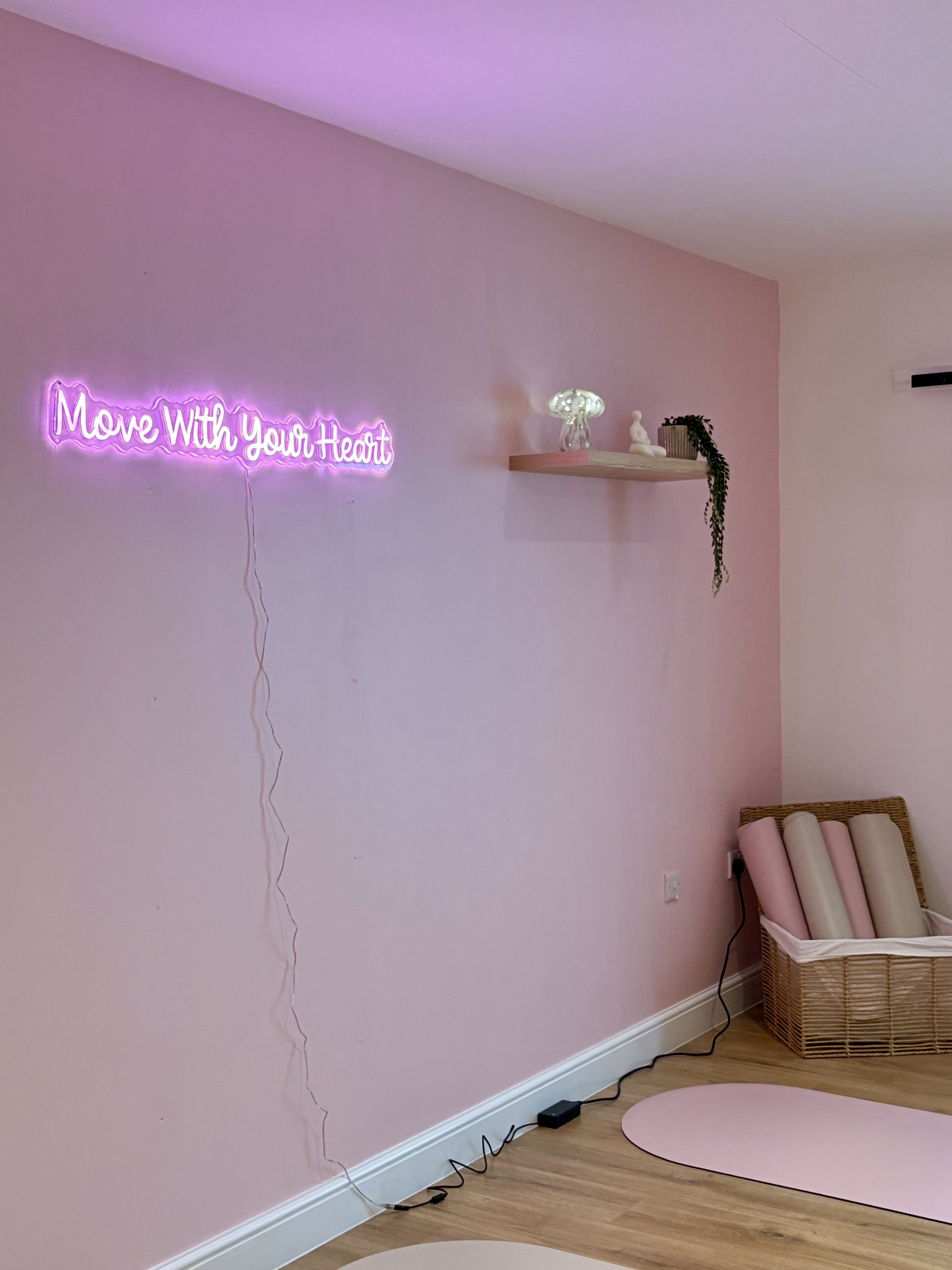Interior of a room with soft pink walls, a neon sign that reads 'Move With Your Heart,' a wooden shelf with decorative objects, a wicker basket with rolled-up yoga mats, and yoga mats on the floor.