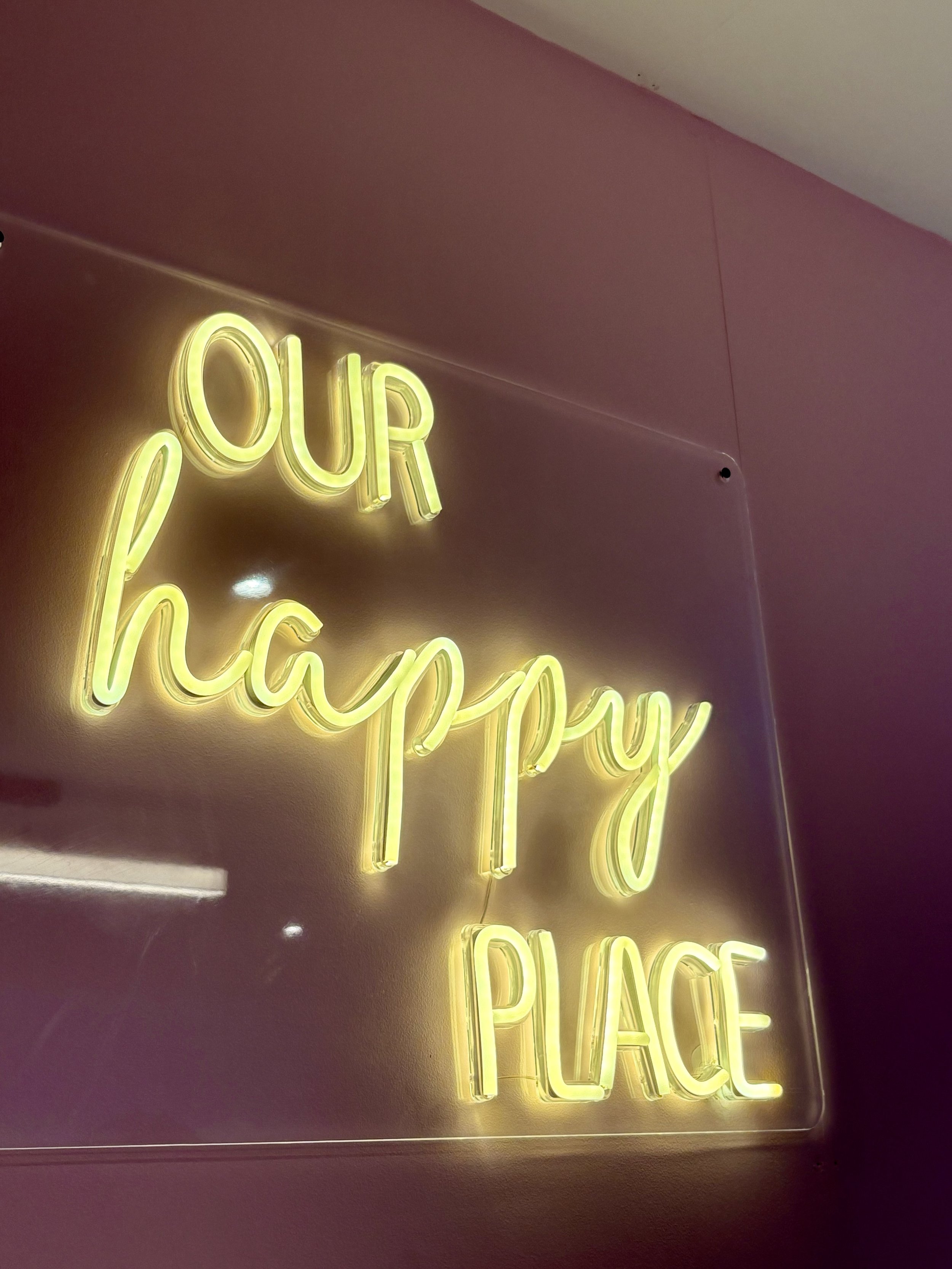 Neon sign reading 'Our Happy Place' in yellow lights on a pink wall.
