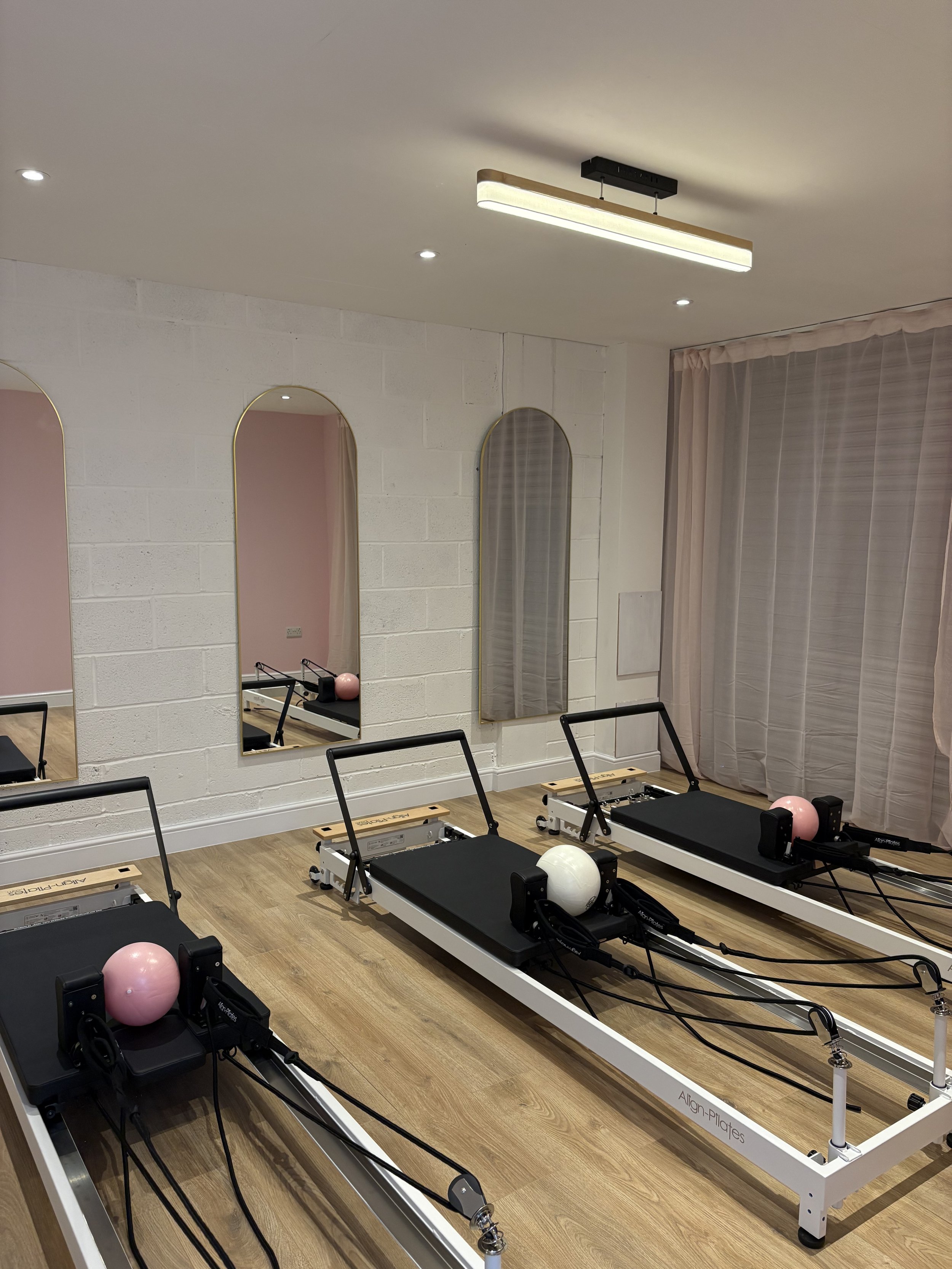 A Pilates studio with three reformer exercise machines, each with a pink or white ball on the foot bar, situated on a wooden floor against a wall with three mirrors and pink curtains.