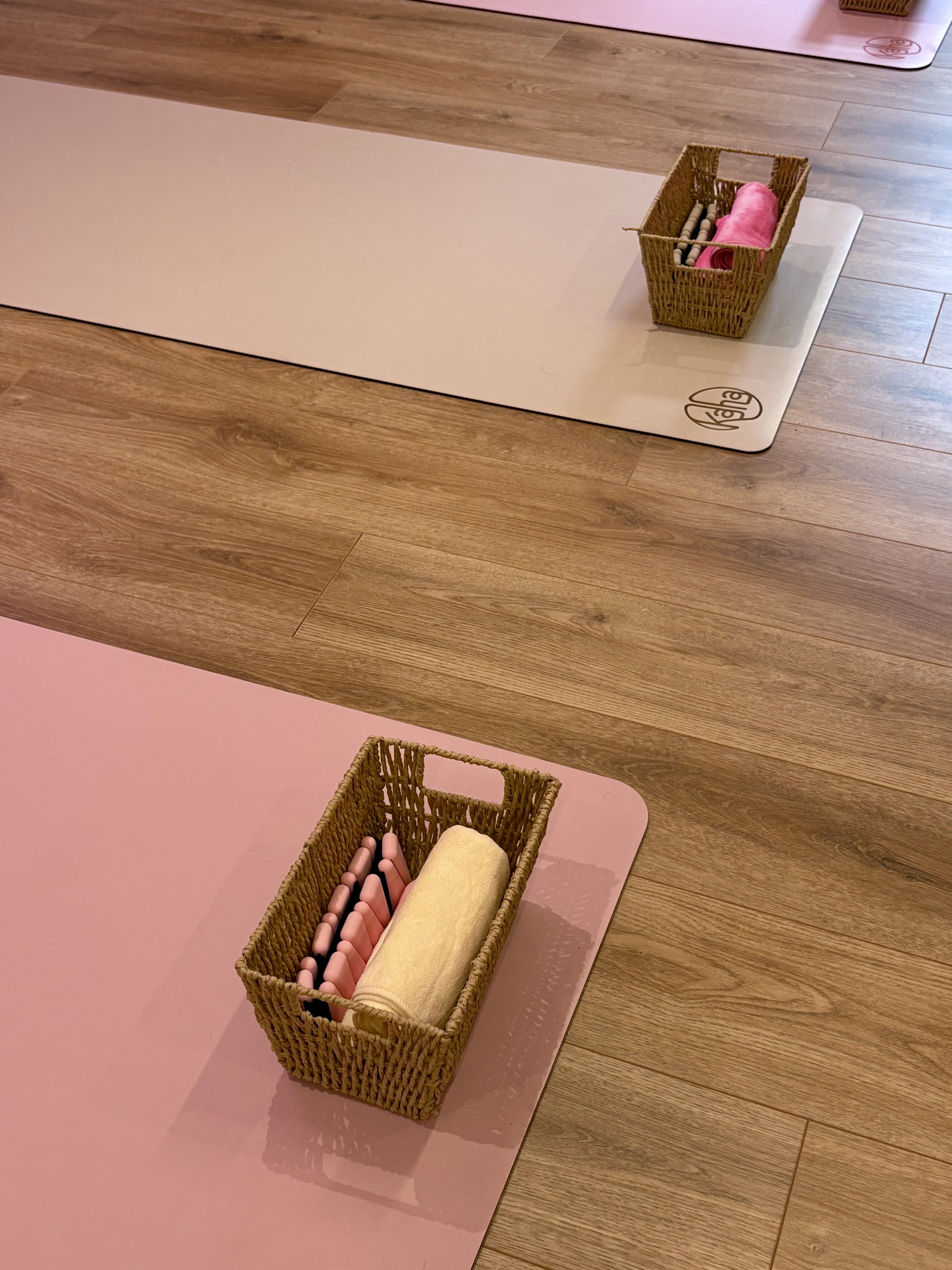 A pink yoga mat with a woven basket containing a rolled yellow towel and several pink yoga blocks, placed on a wooden floor.