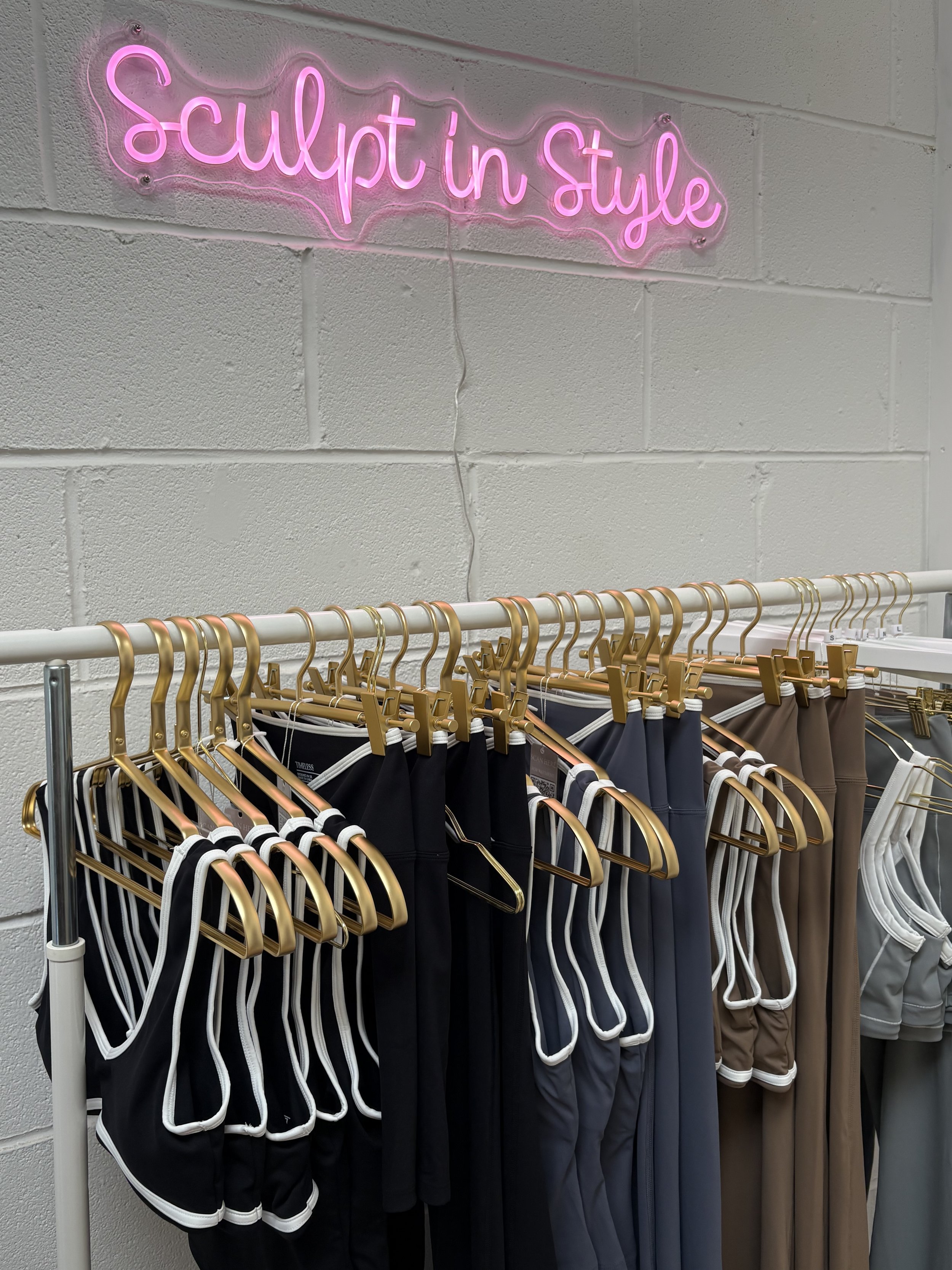 Neon sign reading 'Sculpt in Style' above a clothing rack with athletic shorts in various colors.