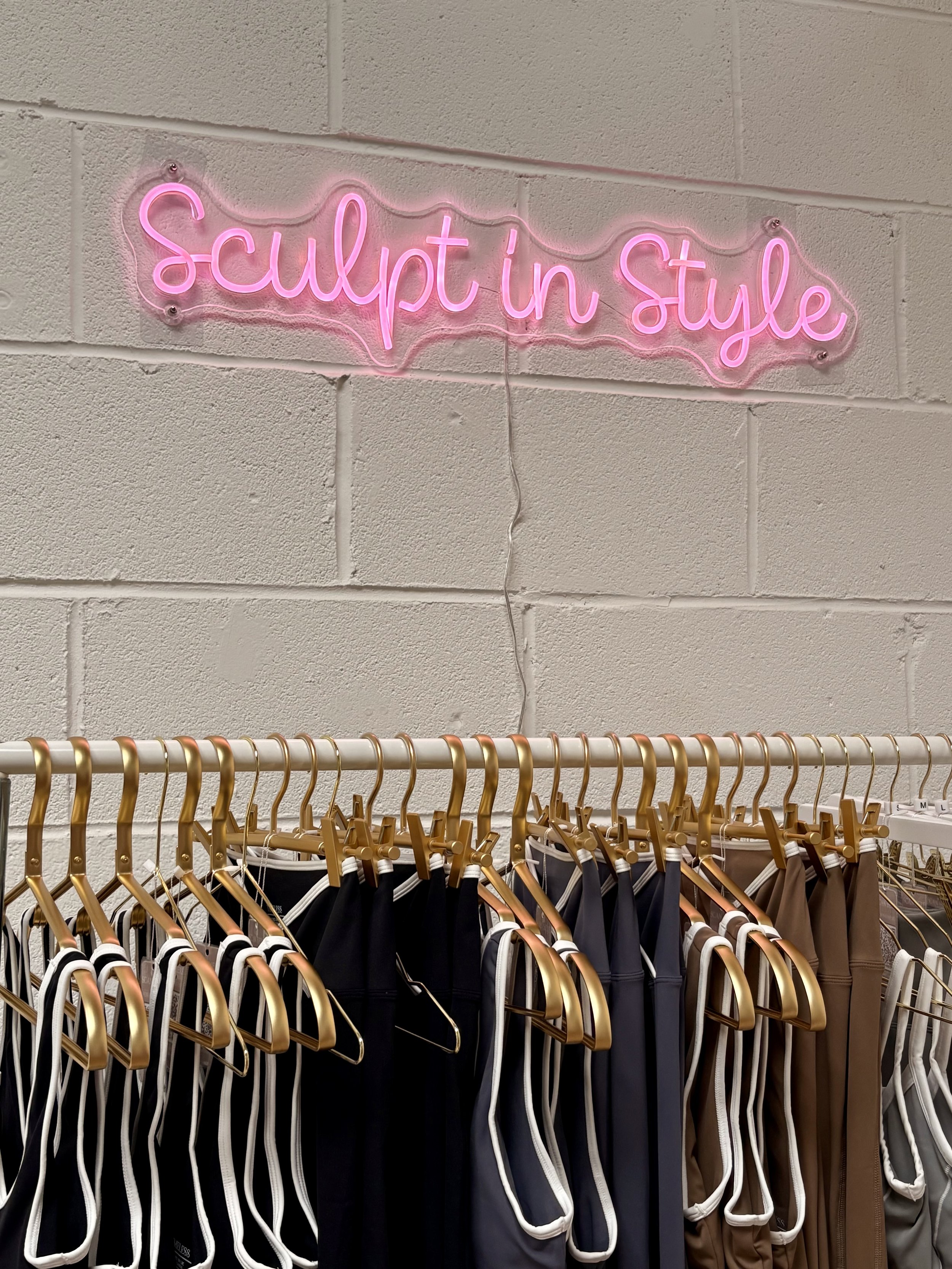 Pink neon sign reading 'sculpt in style' hanging above a clothing rack with various athletic shorts on hangers.