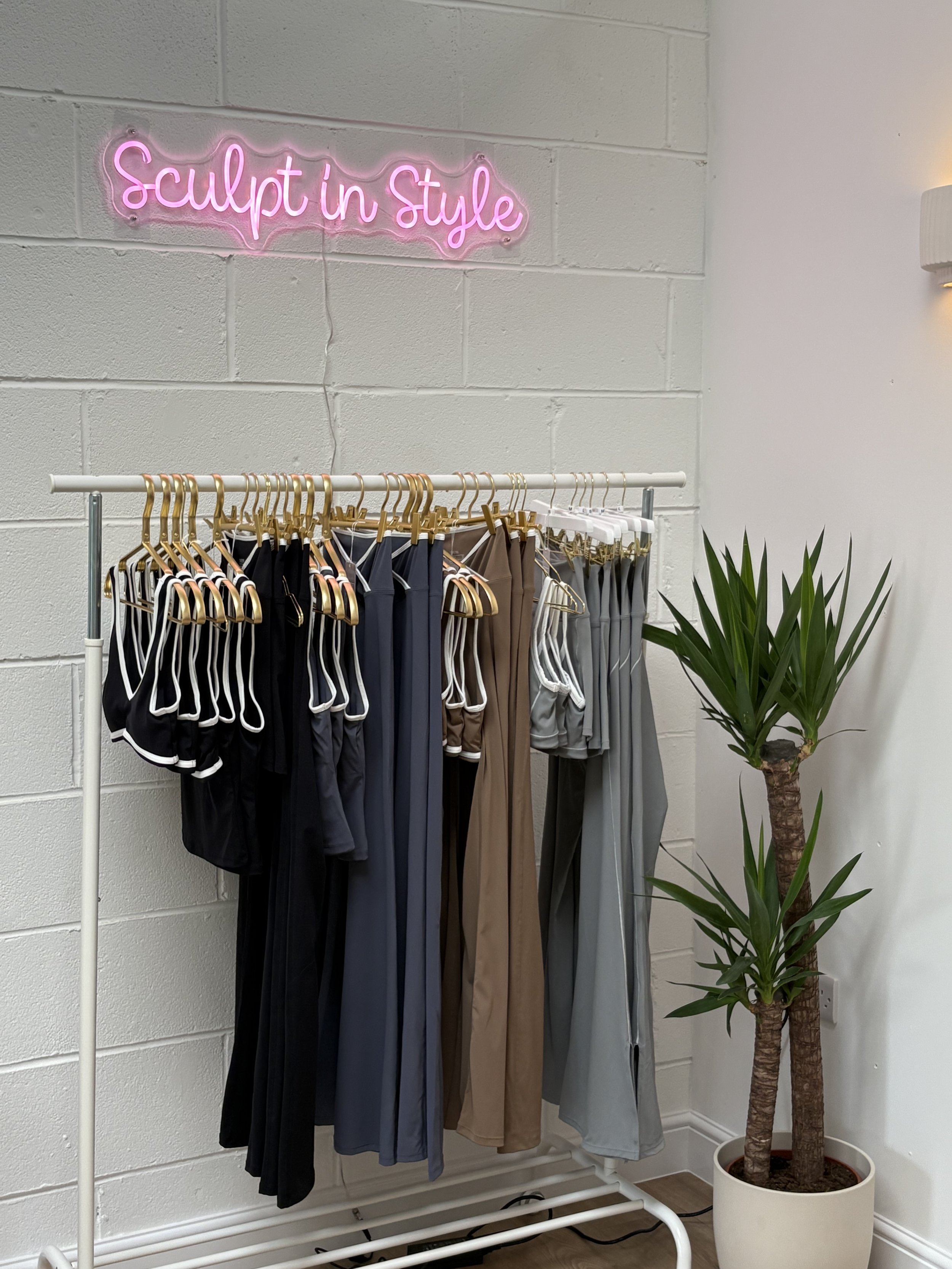 Clothing rack with athleisure wear in black, navy, brown, and gray, next to a potted plant, inside a boutique with a pink neon sign saying 'Sculpt in Style'