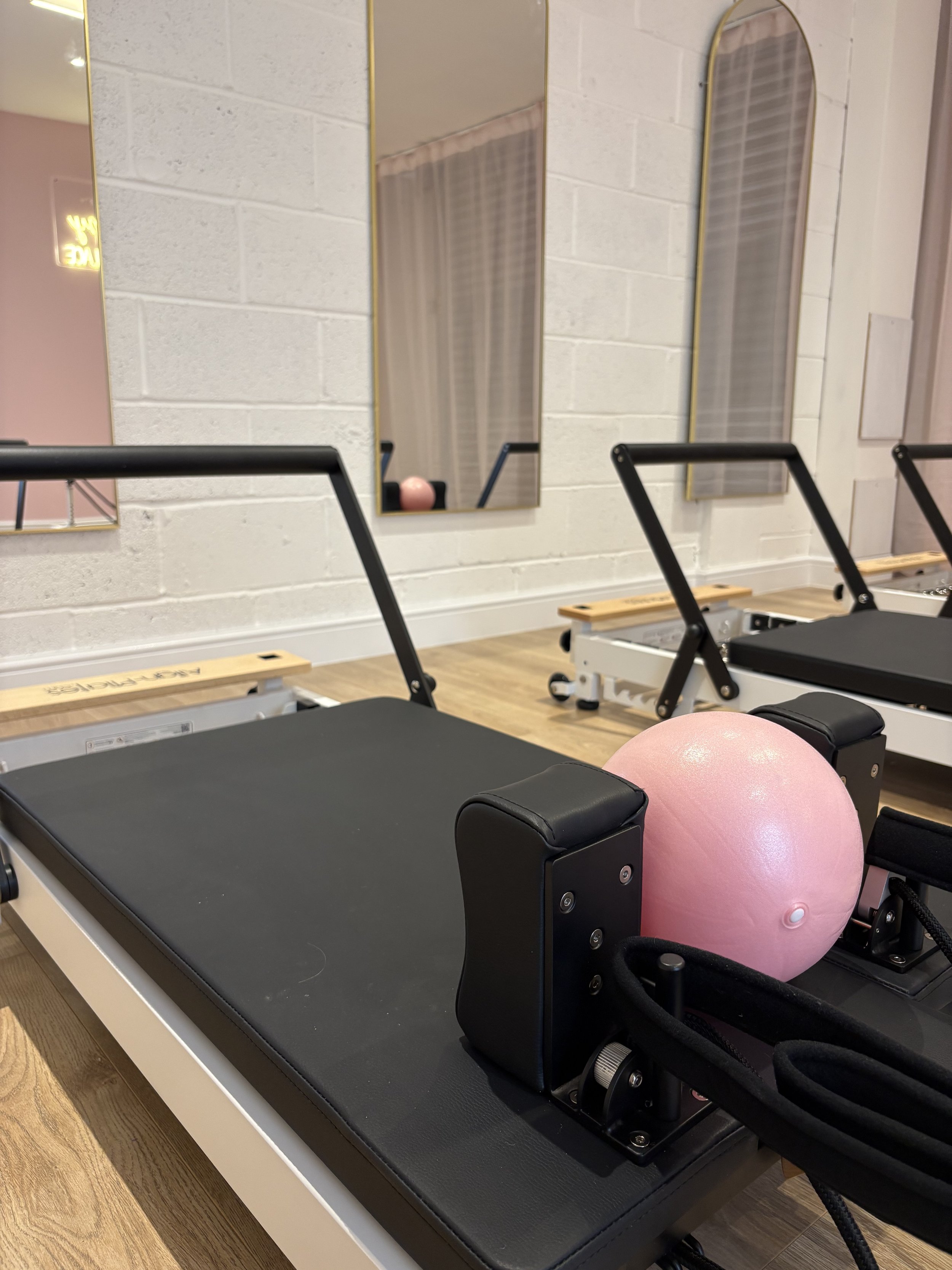 Pilates reformer exercise machine with a pink exercise ball placed on it, in a fitness studio with mirrors on the wall and wooden flooring.