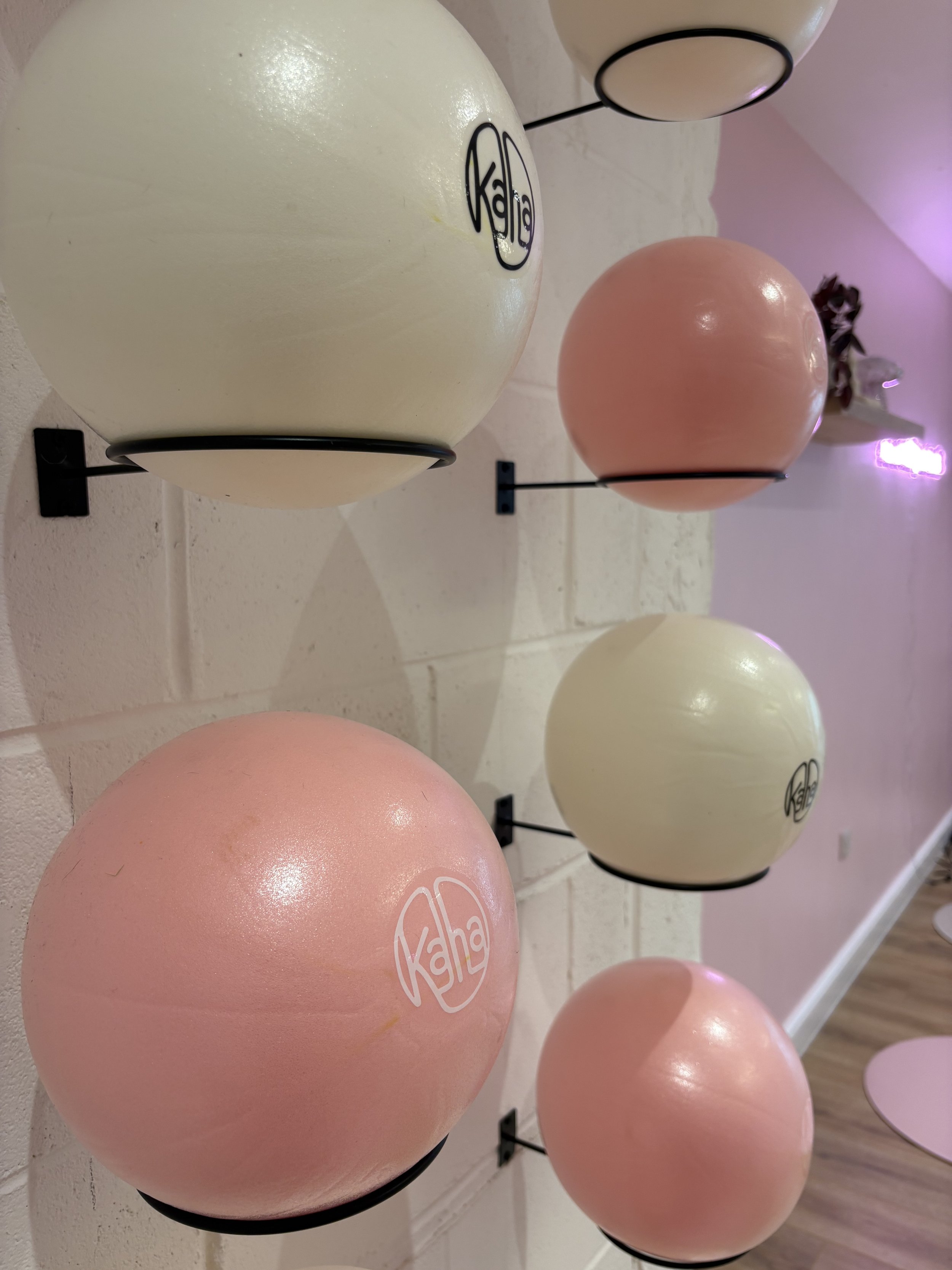 Six spherical objects mounted on black metal brackets on a white brick wall, three are cream-colored and three are pink, with the word 'Kaha' printed on them.