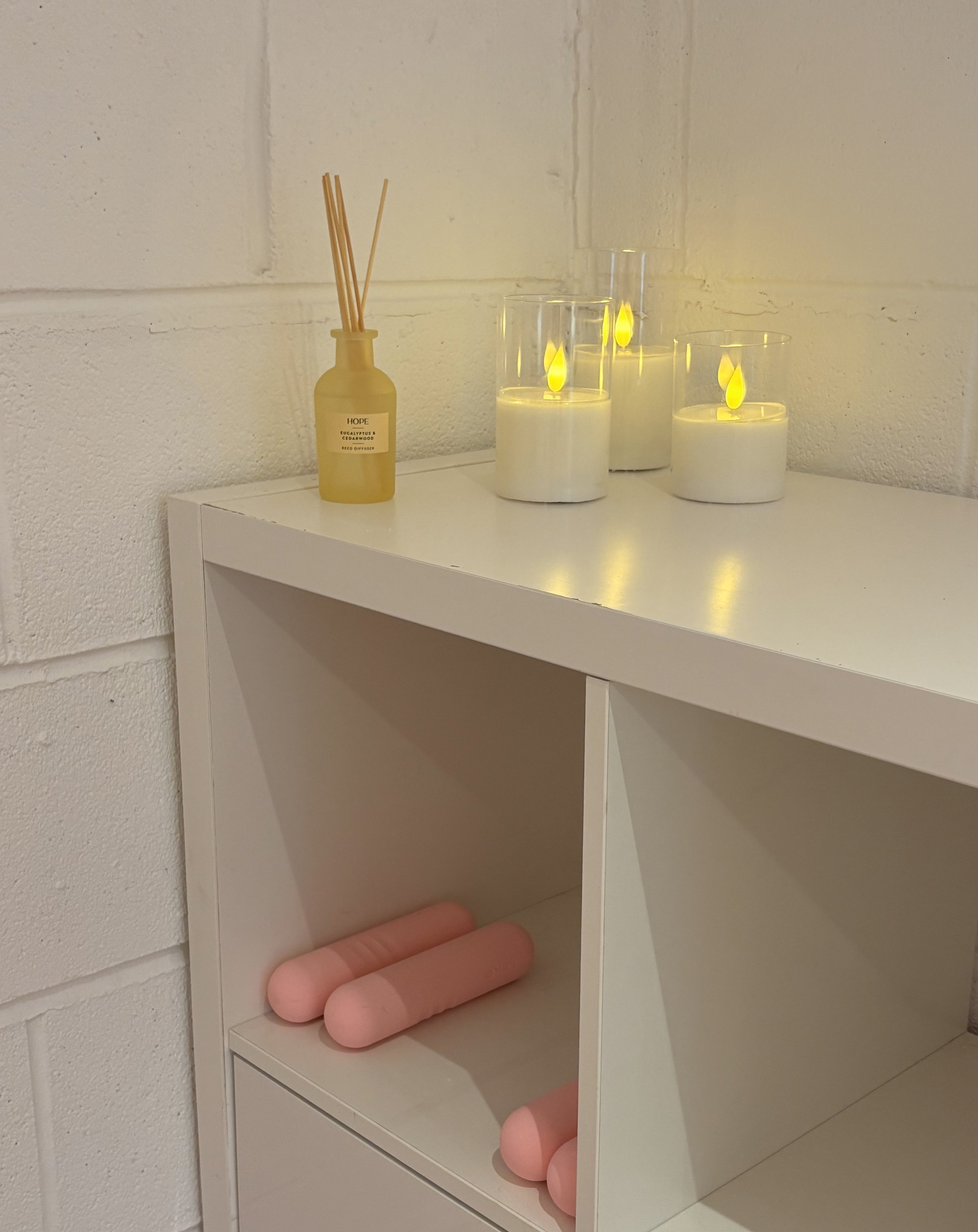 A white shelf with pink foam rollers in a lower compartment and three candles with yellow flickering flame-shaped LED lights on top. A yellow reed diffuser bottle labeled 'HOPE' with reed sticks is also on the top surface against a white brick wall.