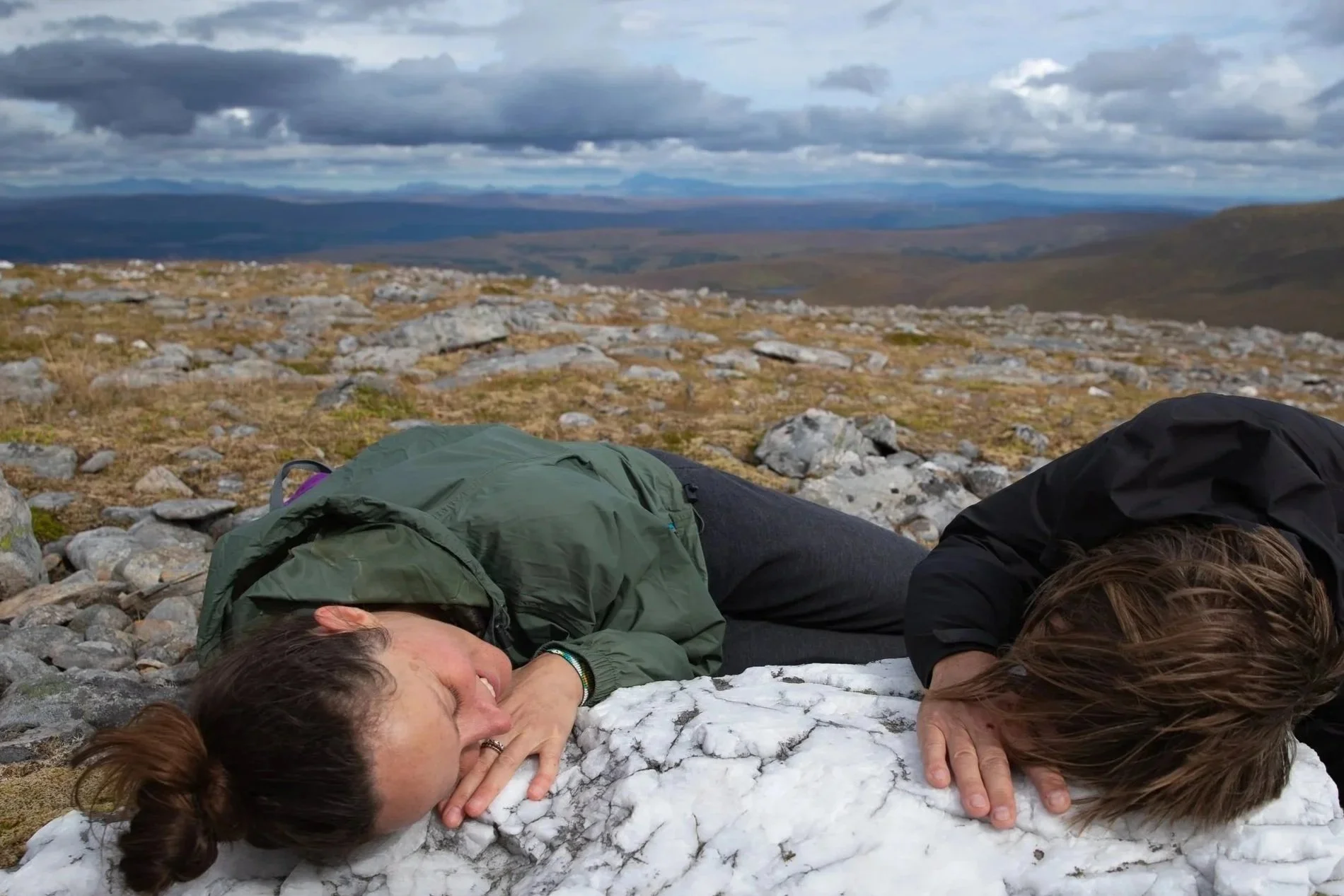 Two women lying on rocky ground, resting their heads on a large white stone, with open landscape and mountains in the distance under cloudy sky.
