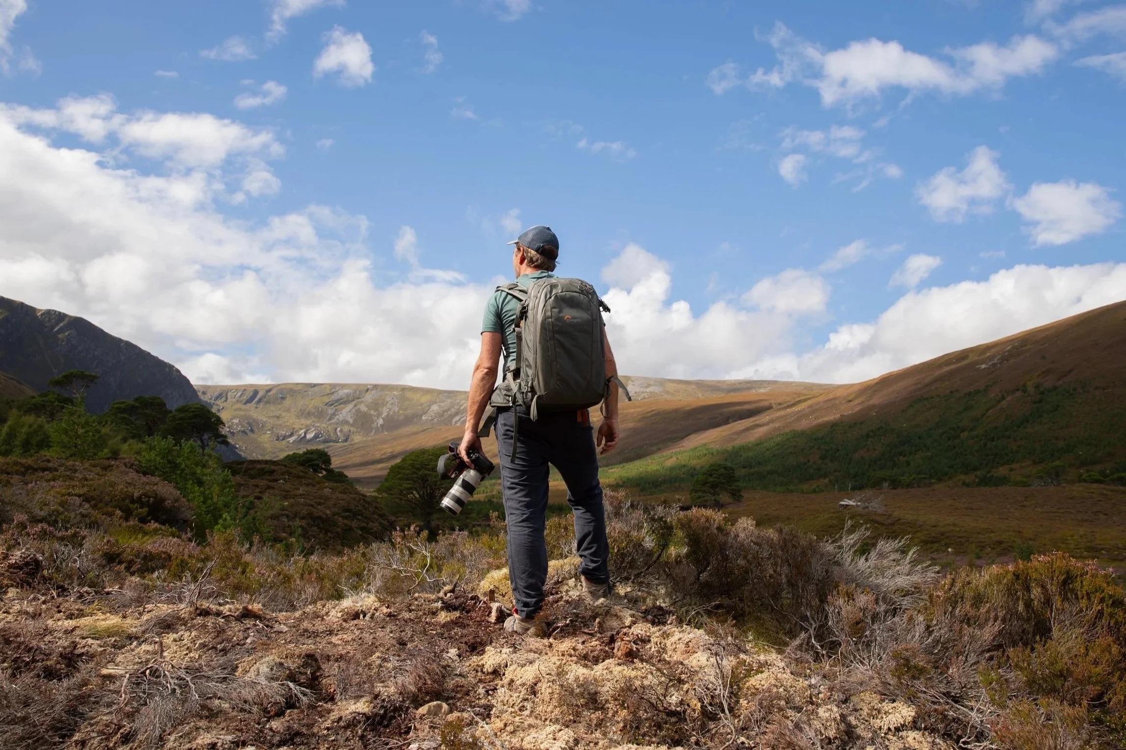 A man with a backpack holding a camera stands on a rocky trail in a hilly landscape with sparse trees and green shrubbery, under a partly cloudy sky.