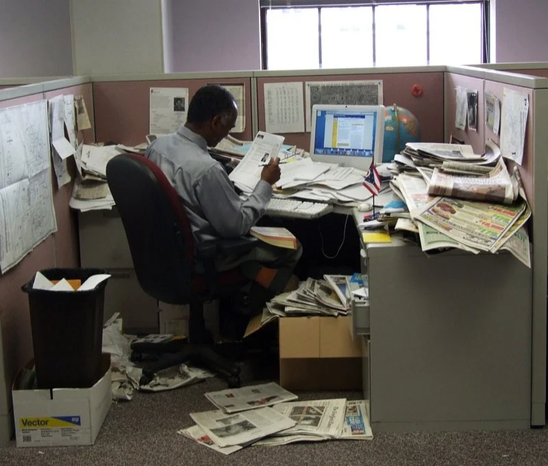 Person sitting in cluttered office cubicle surrounded by piles of papers and mess.