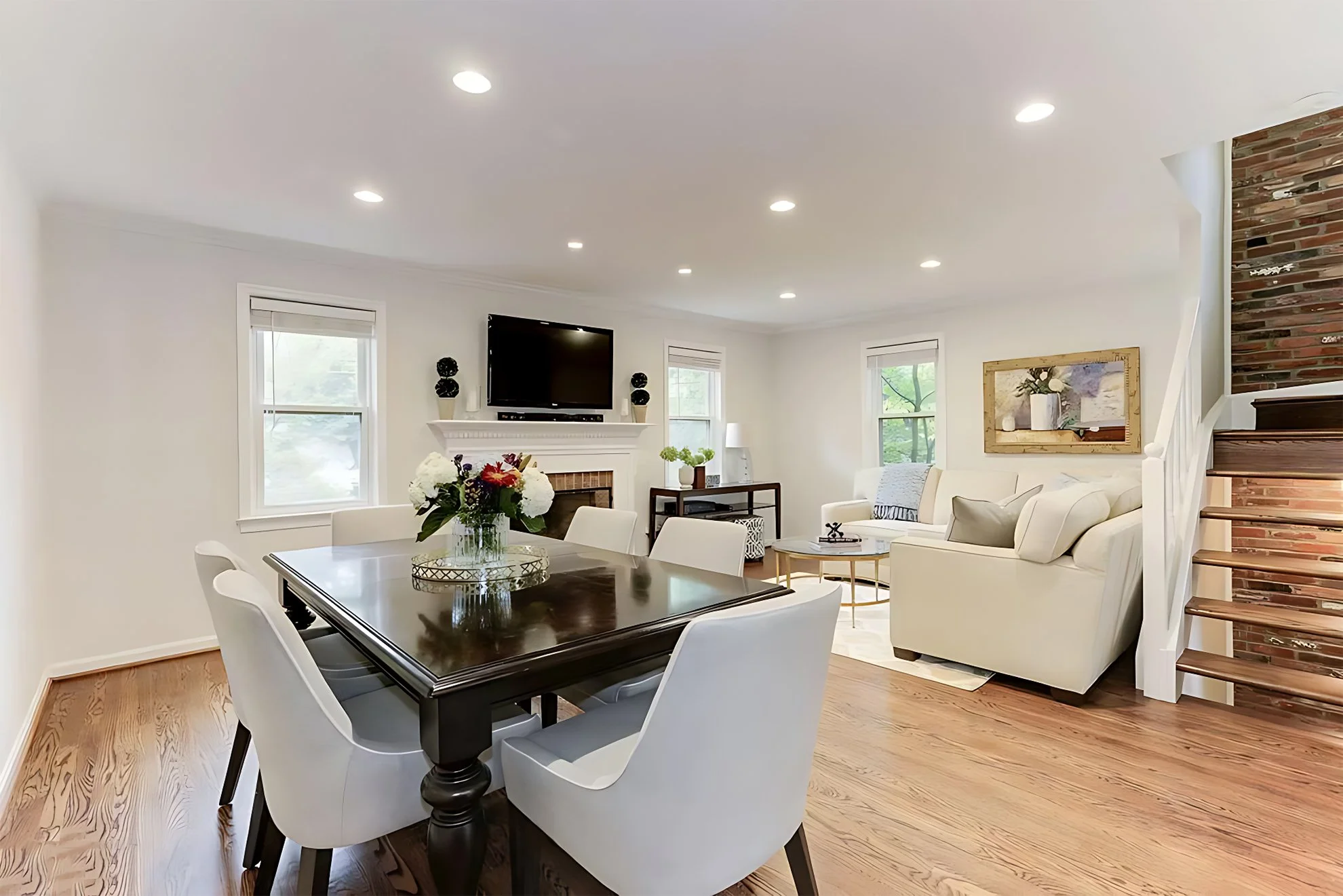Bright dining and living area with wooden floors, white chairs, and fireplace in the background.