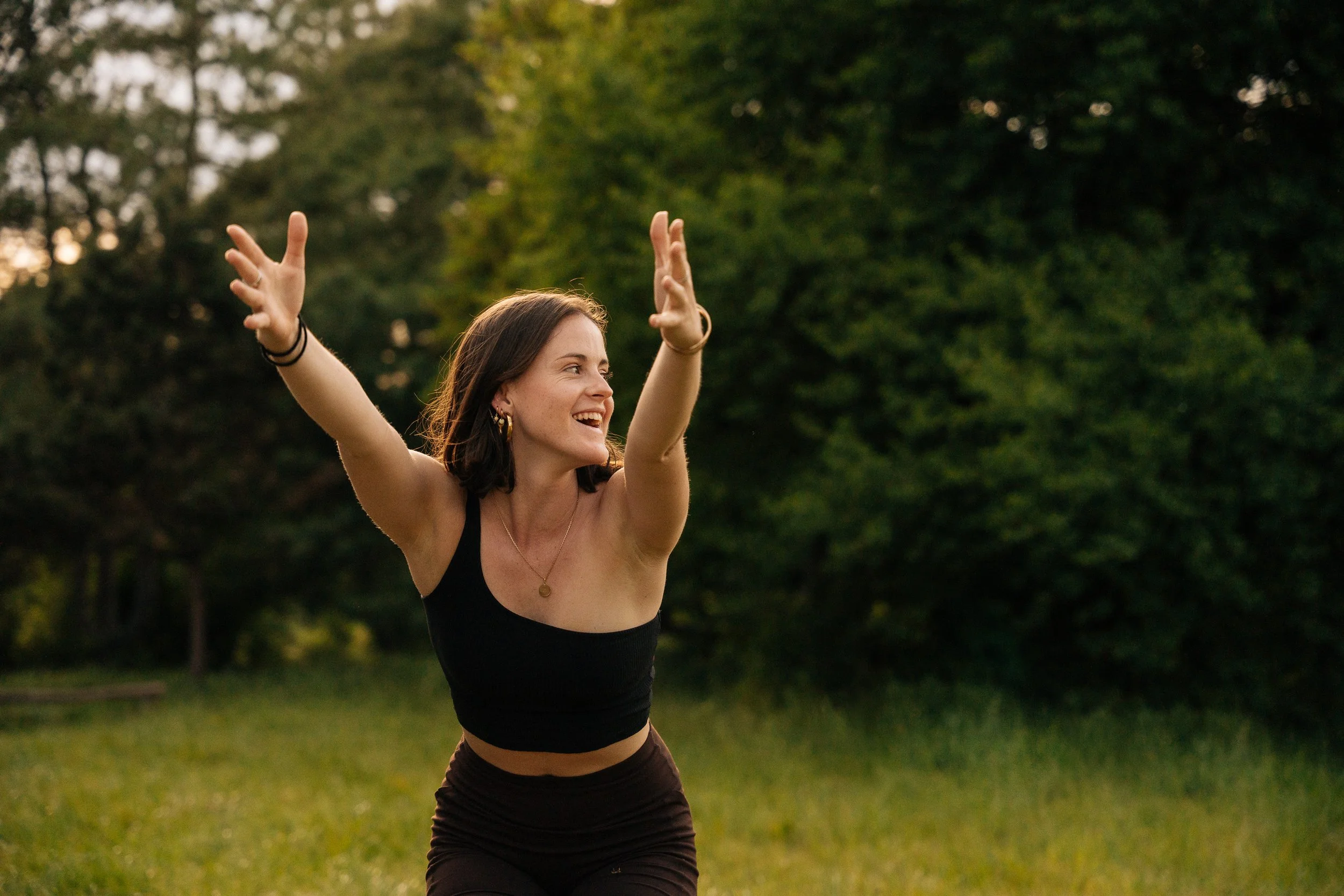 Mathilde Lejour. Une femme souriante déployant ses bras dans un parc verdoyant au coucher du soleil.