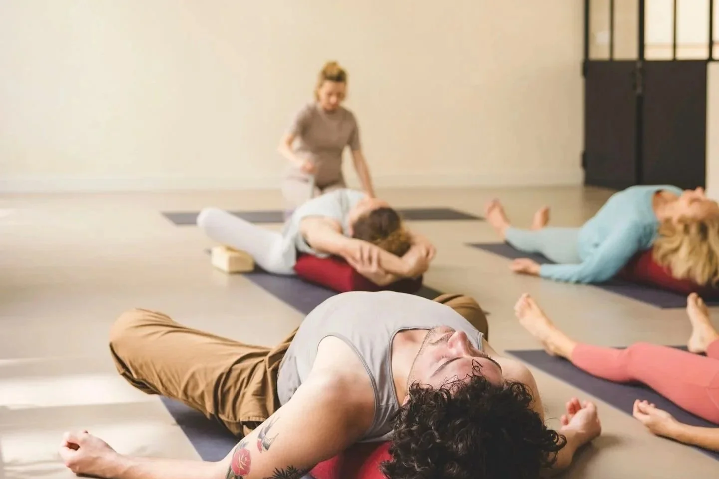Formation Yin Yoga 50h module 2, approfondir son enseignement. Les élèves sont allongés sur un bolster en savasana. La professeure est en arrière plan et guide la relaxation en jouant un bol de cristal. Les couleurs sont douces et naturelles.