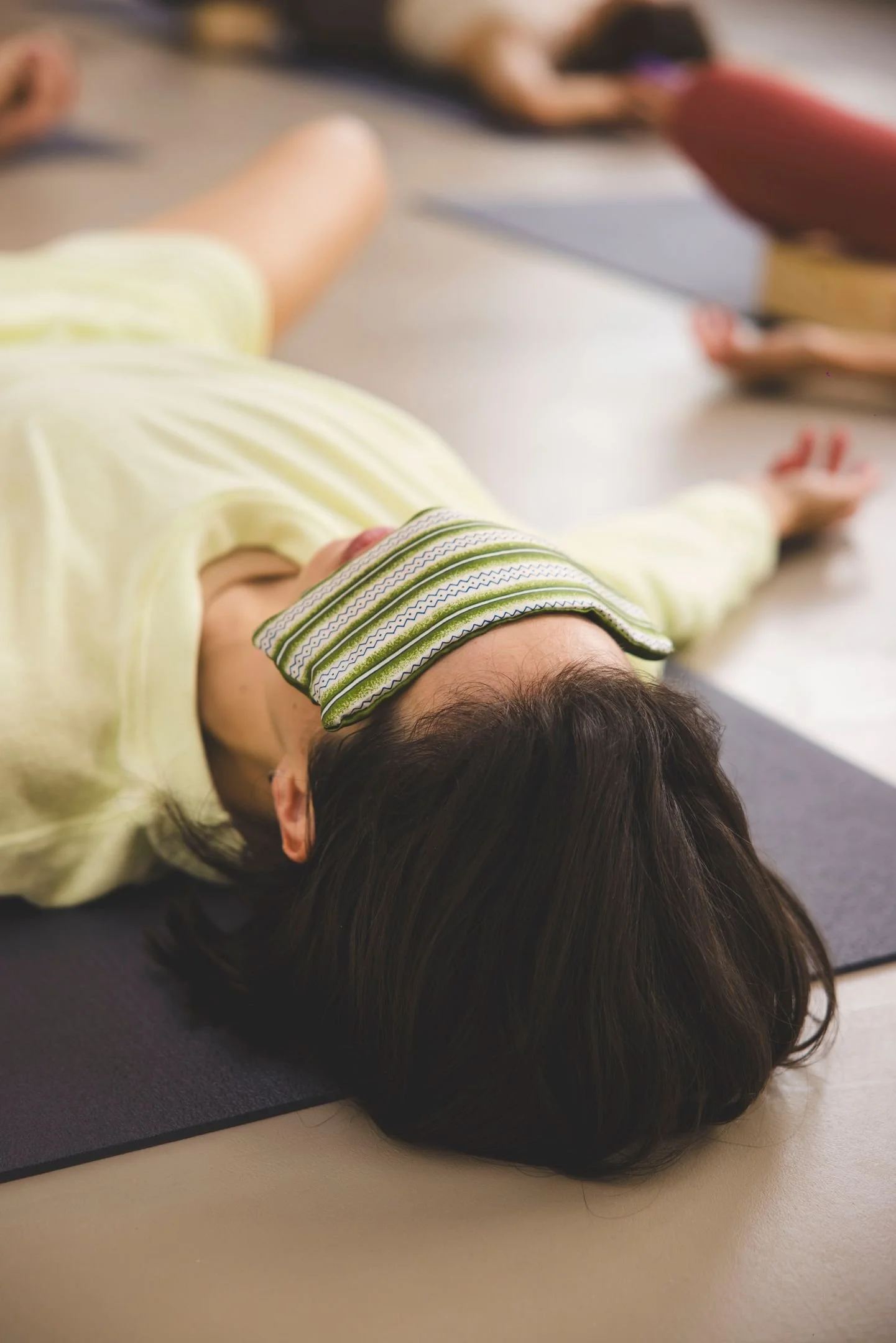 Eleve de yoga allongée sur son tapis avec un masque sur les yeux, en train de se détendre en savasana. Elle porte un tee-shirt jaune clair et le masque pour les yeux est en tissu vert avec des motifs géométriques.