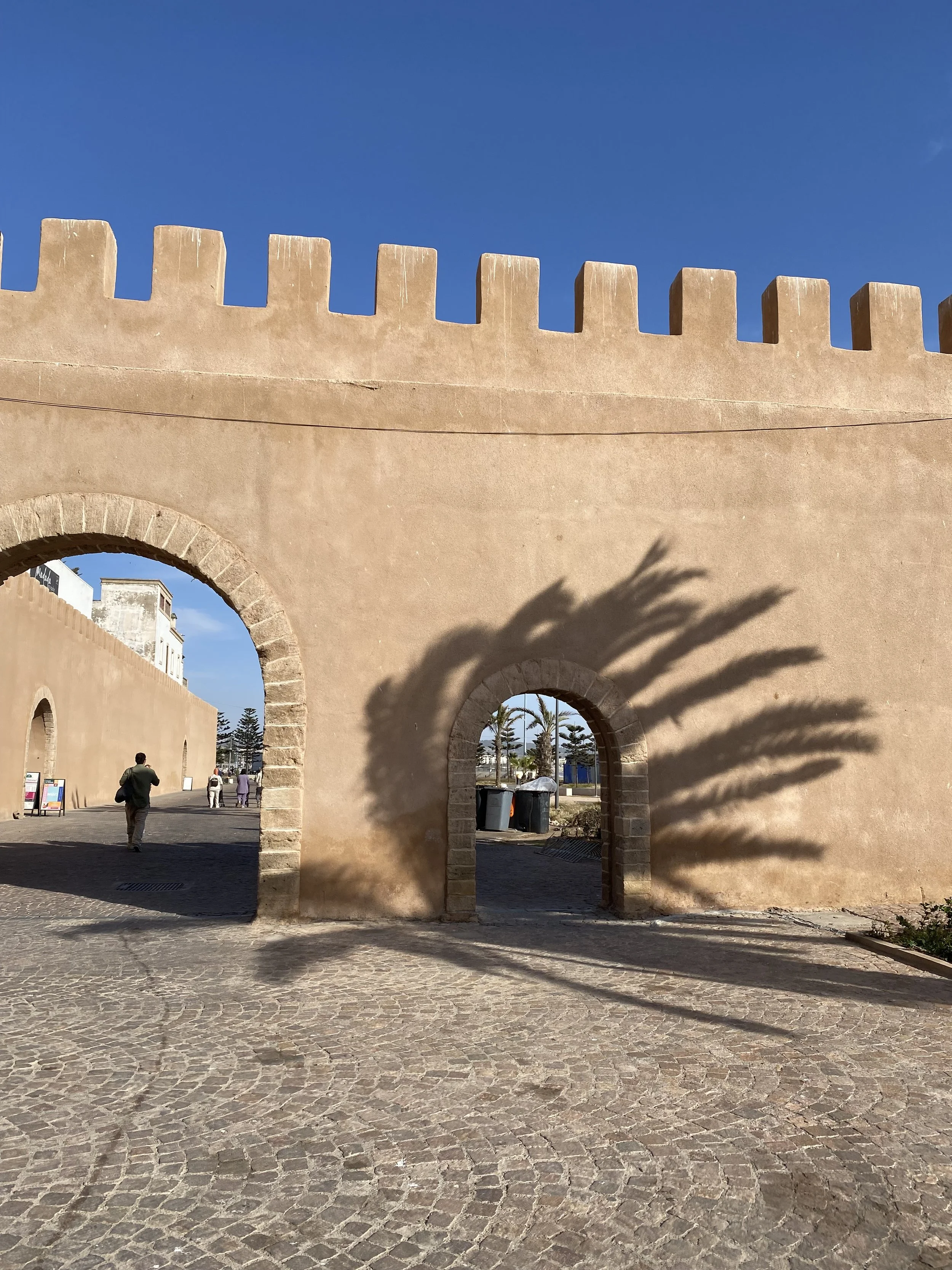 Mur en pierre avec créneaux et arcs, ombre de palmier projetée, ciel bleu en arrière-plan.